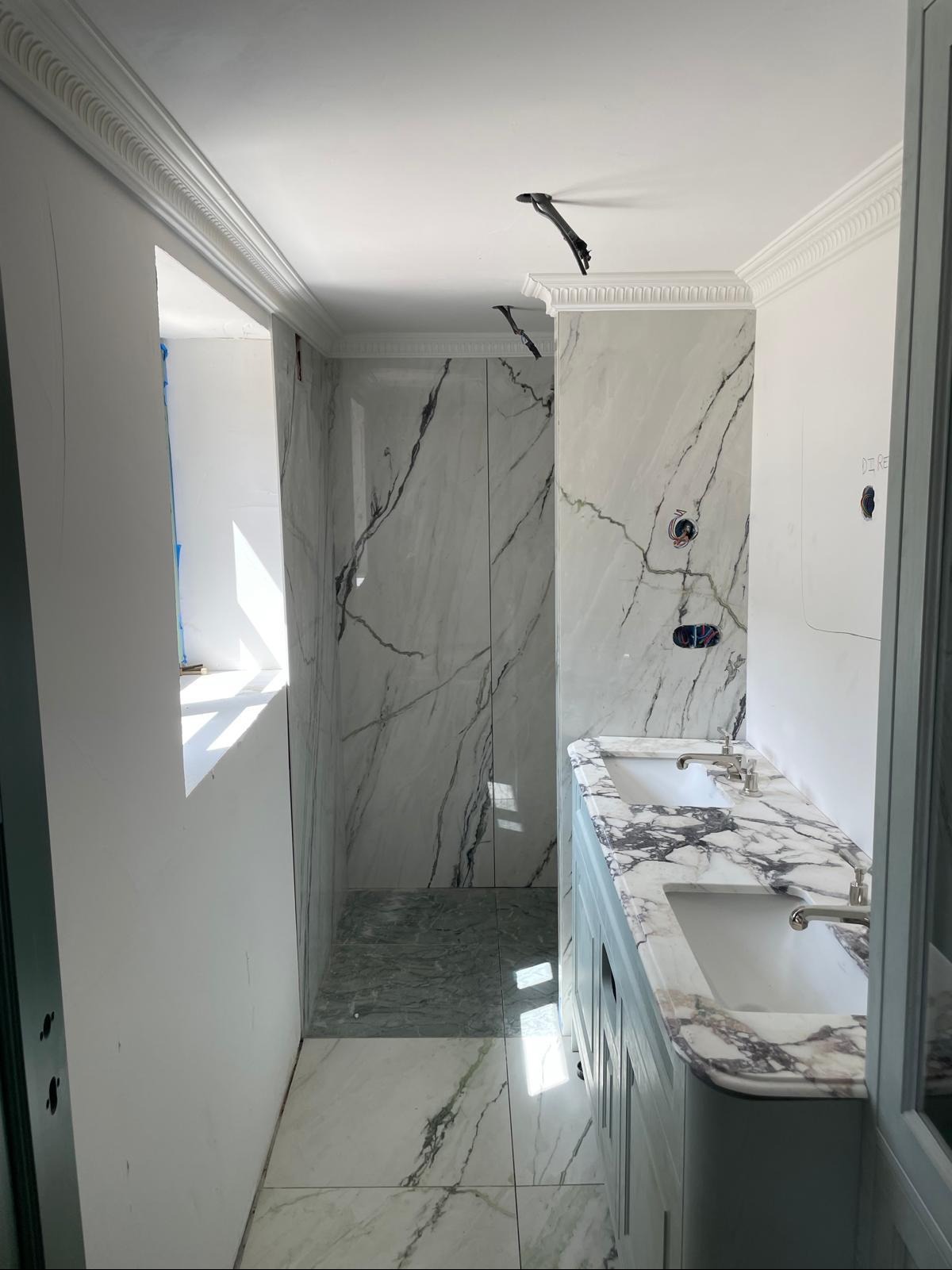 Wykończona łazienka z marmurowymi płytkami na ścianach i podłodze, podwójna umywalka z marmurowym blatem, dekoracyjne listwy sufitowe, surowy stan ścian w tle.