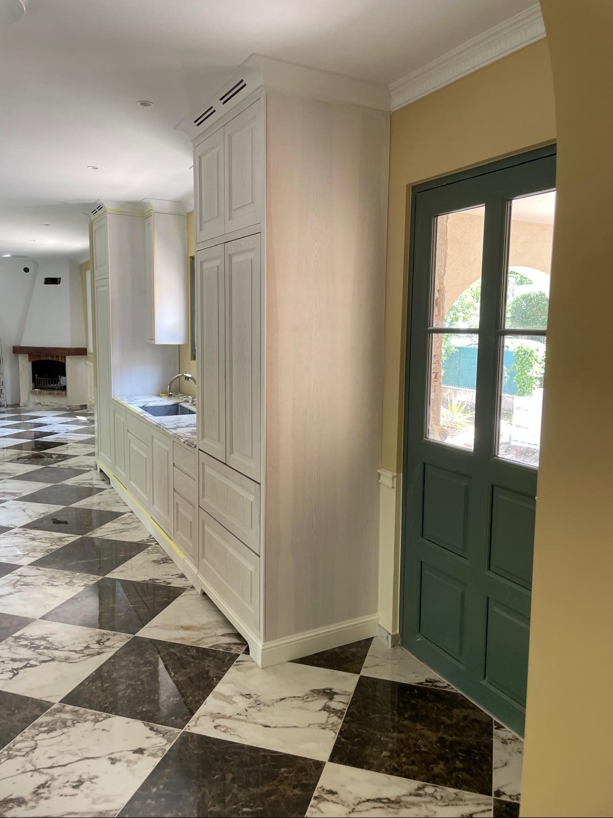 Jasna zabudowa kuchenna z marmurowym blatem i wzorzystą podłogą w odcieniach szarości, widok z perspektywy wejścia, z zielonymi drzwiami po prawej stronie.