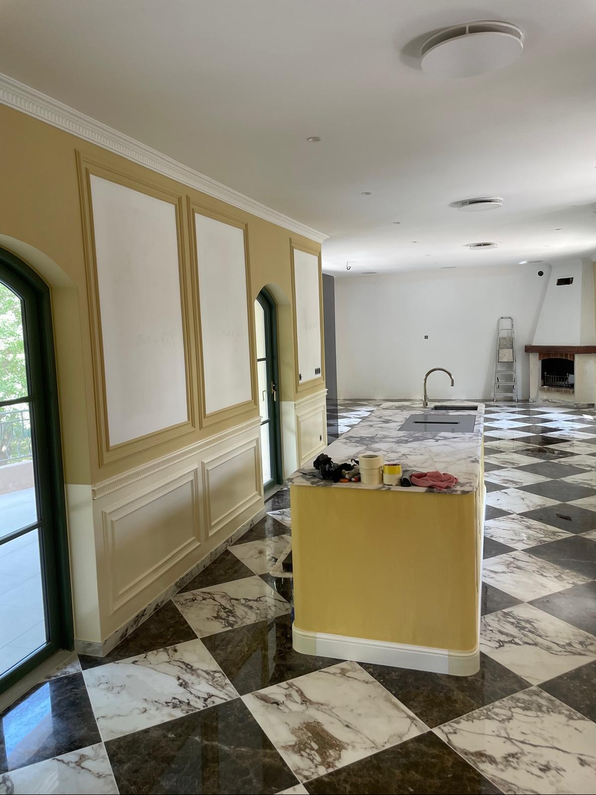 Przestronne wnętrze w trakcie wykończenia: żółte ściany z białymi panelami, marmurowa podłoga w szachownicę, wyspa kuchenna i kominek w tle. Widoczne narzędzia malarskie.