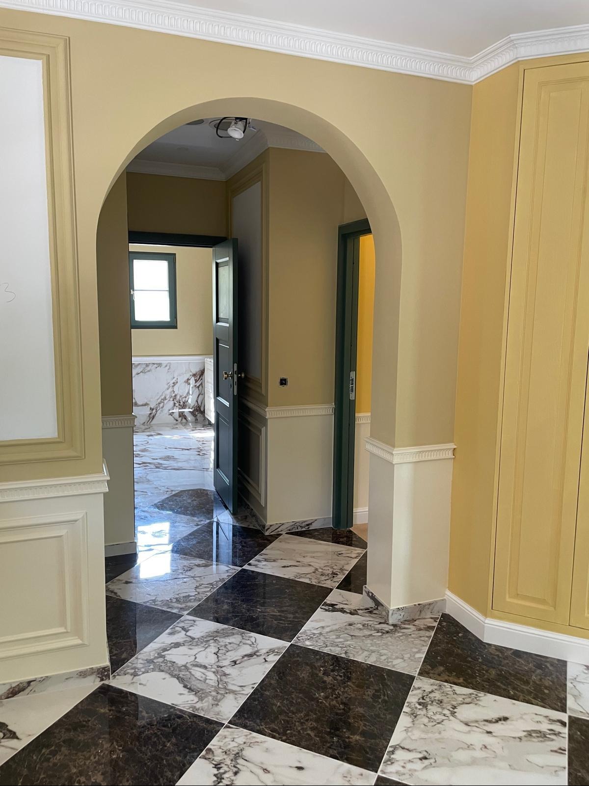 Eleganckie wnętrze z marmurową podłogą w szachownicę, łukowatym przejściem i żółtymi ścianami. Widoczne detale sztukaterii i otwarte drzwi do kolejnych pomieszczeń.