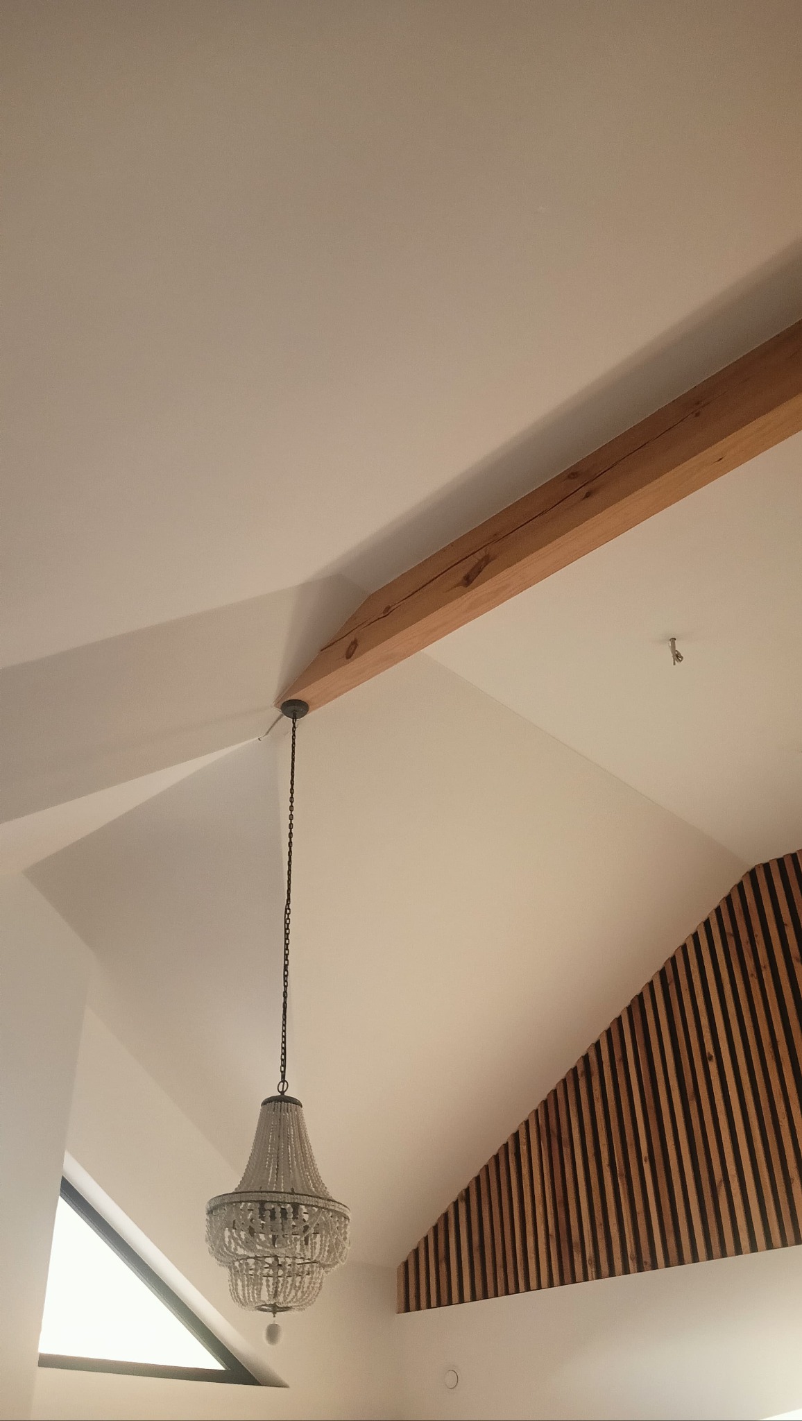 Biały sufit ze skośną ścianą, drewnianą belką i dekoracyjną listwą. Kryształowy żyrandol zwisa na łańcuchu. Widoczny fragment okna.