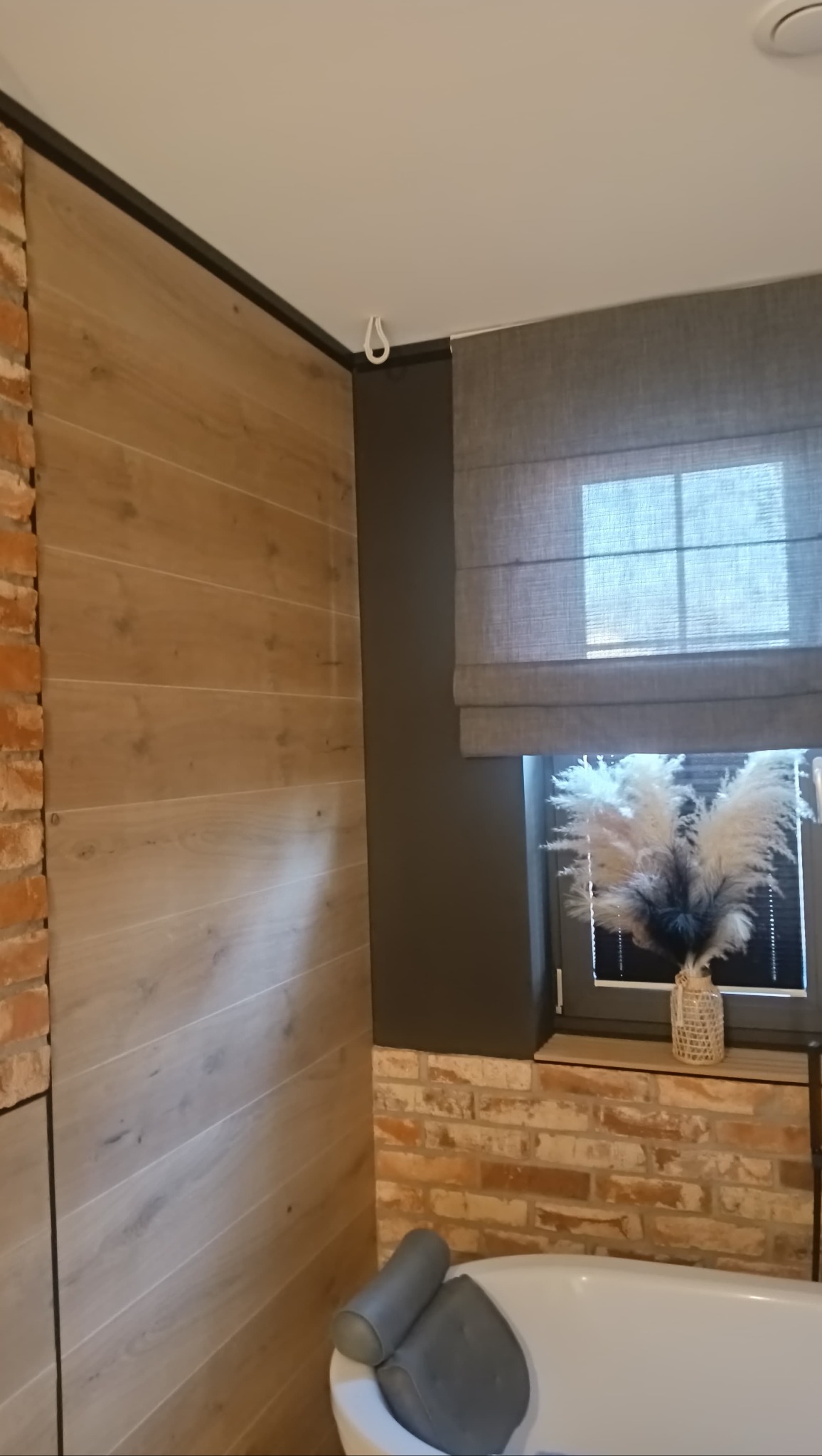 Nowoczesna łazienka z akcentami drewna i cegły: drewnopodobne panele ścienne, ceglana ściana, szara roleta rzymska i wanna z zagłówkiem. Minimalistyczny design.