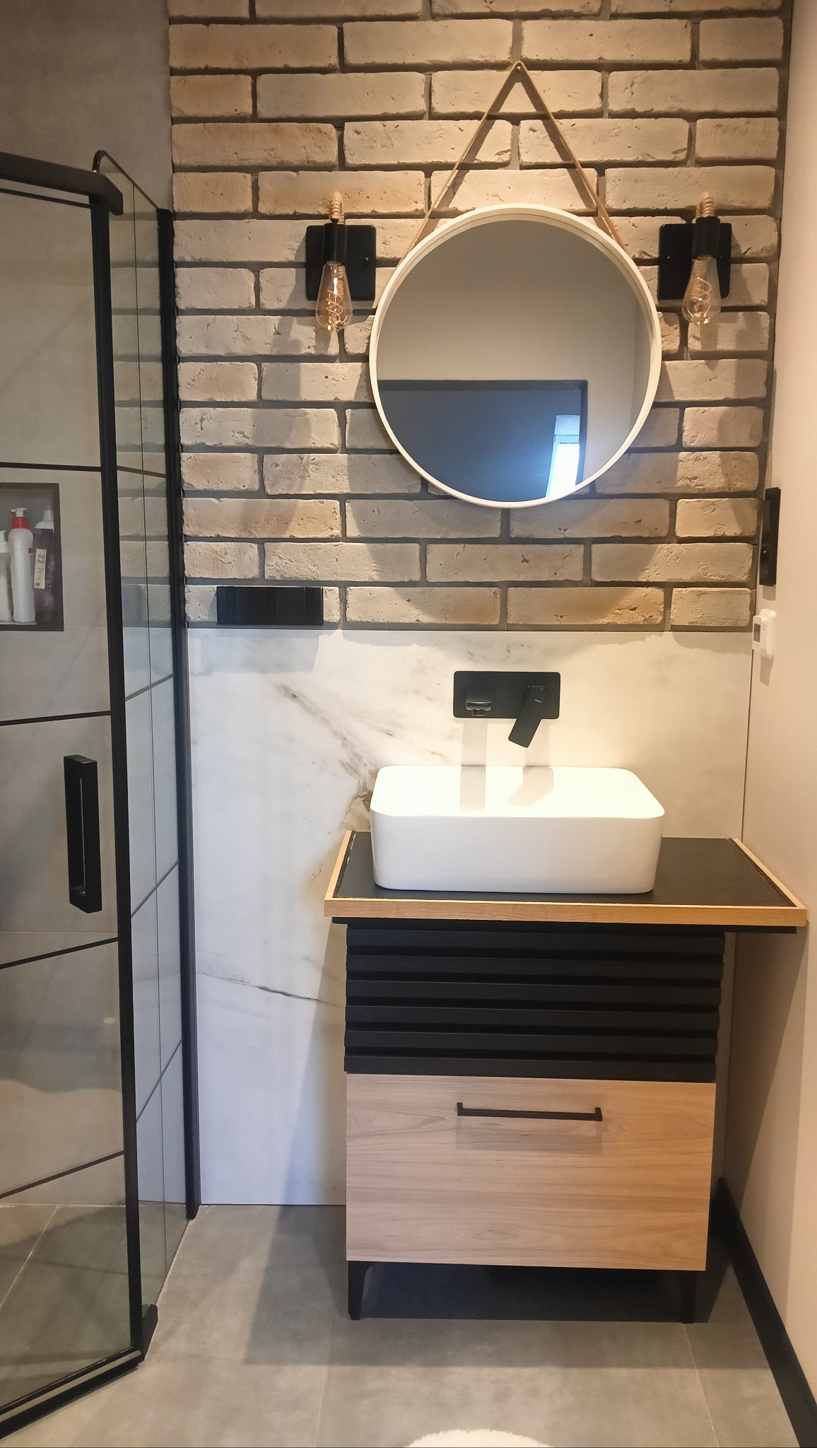 Nowoczesna łazienka z umywalką nablatową, lustrem na ceglanej ścianie i kabiną prysznicową. Minimalistyczny design z elementami loftowymi.