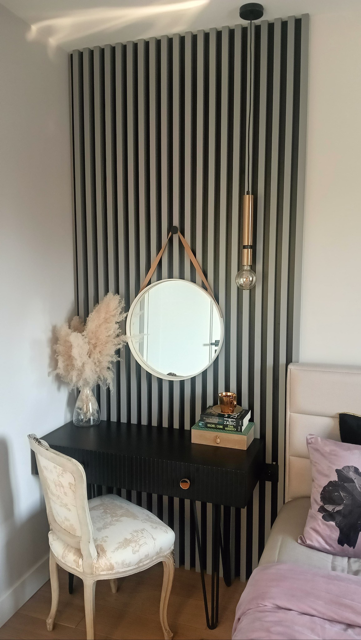 Stylowa aranżacja kącika z toaletką: czarne biurko z lustrem na skórzanym pasku, dekoracyjna ściana z lameli, designerska lampa i krzesło w stylu vintage.