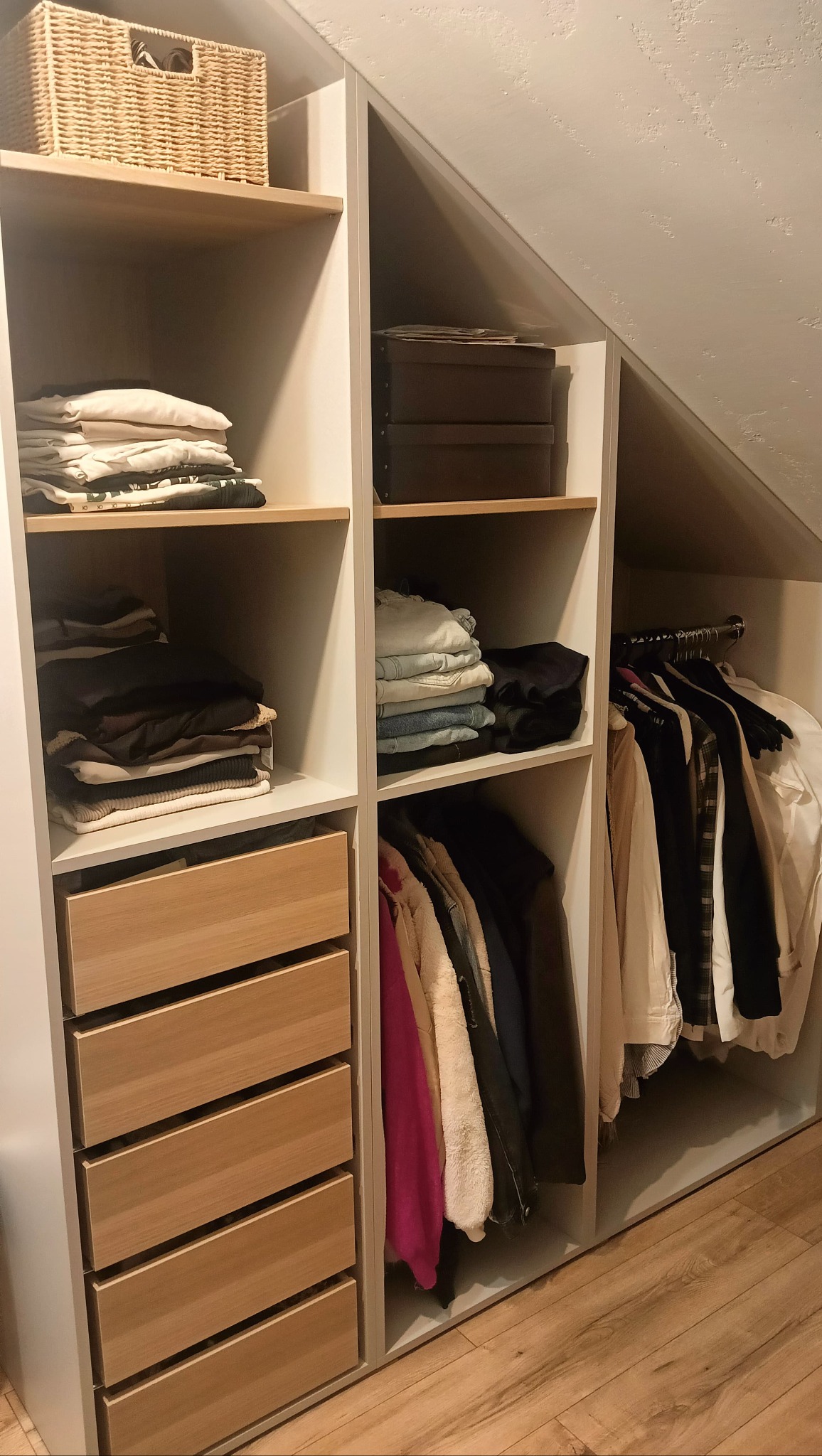 Biała szafa wnękowa z drewnianymi szufladami i półkami, wypełniona ubraniami i koszem, dopasowana do skosu dachu, na drewnianej podłodze.