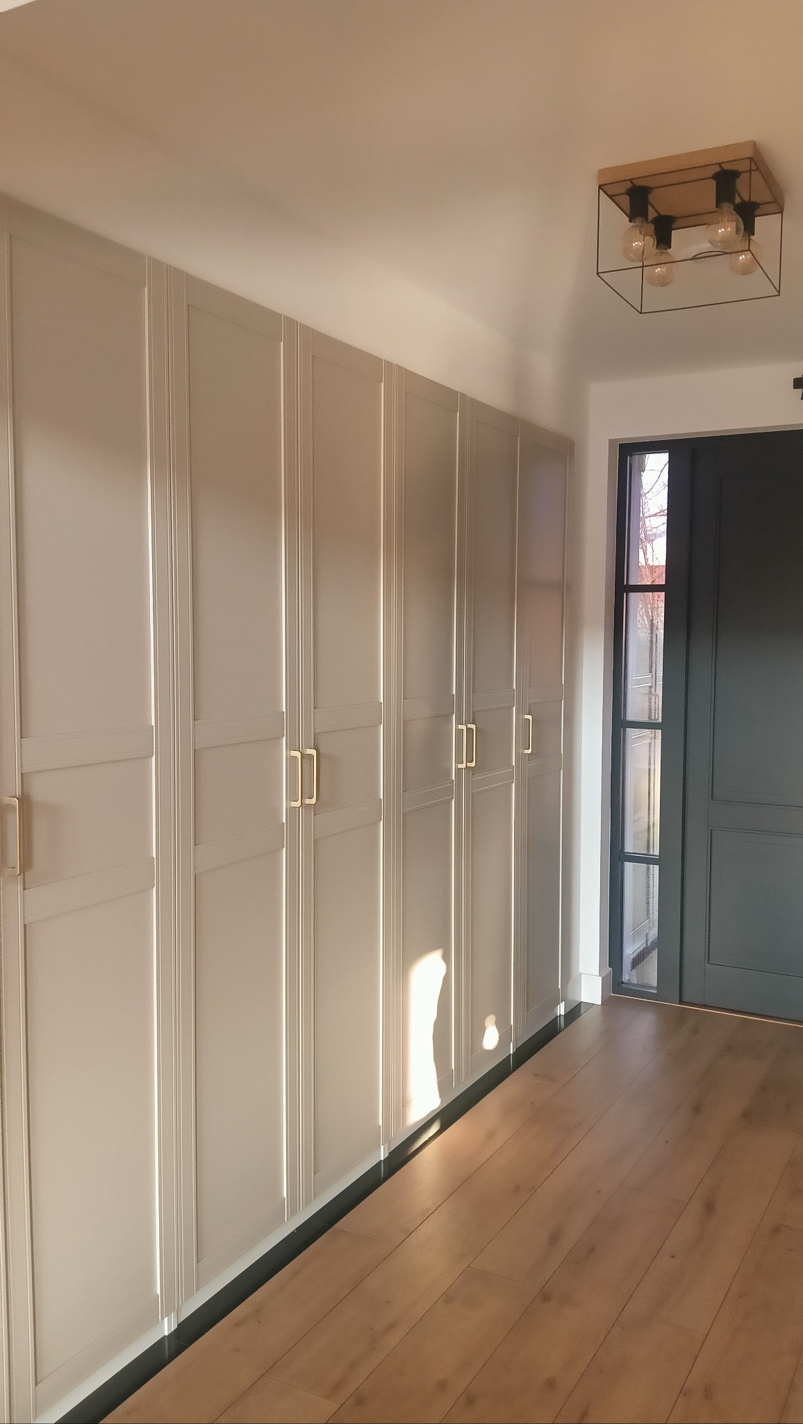 Biała zabudowa szafy wnękowej z frezowanymi frontami i złotymi uchwytami, drewniana podłoga, nowoczesna lampa sufitowa, szara ściana i drzwi wejściowe.