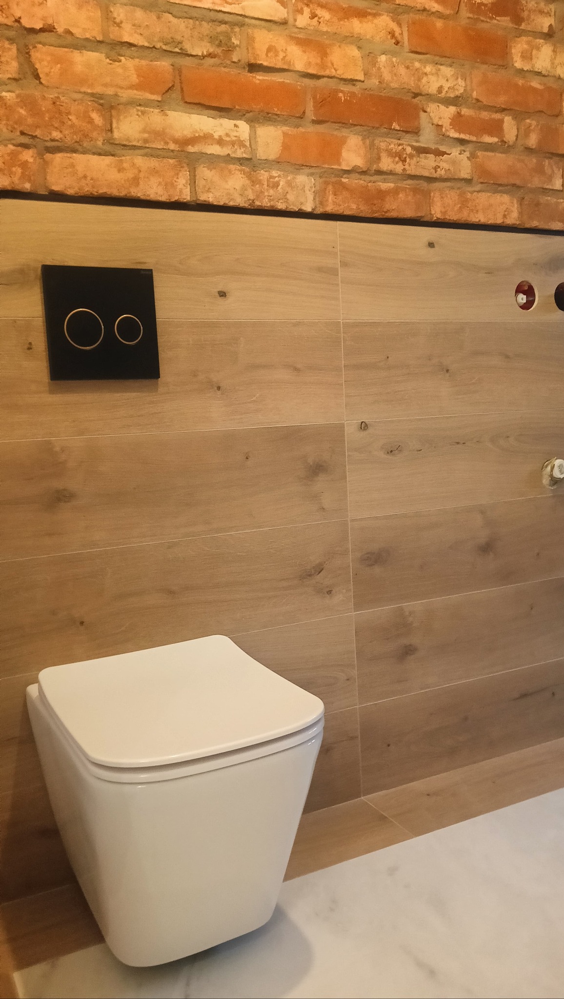 Wisząca toaleta na tle drewnopodobnych płytek i ceglanej ściany. Czarny panel spłuczki z dwoma okrągłymi przyciskami. Widoczne elementy instalacji.