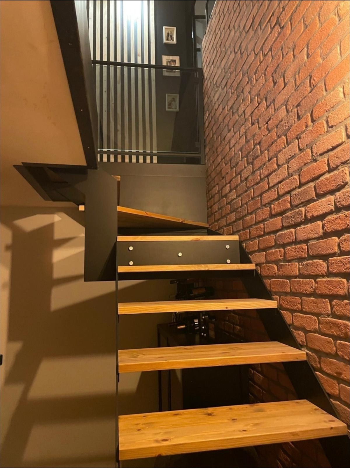 Nowoczesne schody z drewnianymi stopniami i metalową konstrukcją, przylegające do ściany z czerwonej cegły. Widoczne elementy dekoracyjne i oświetlenie.