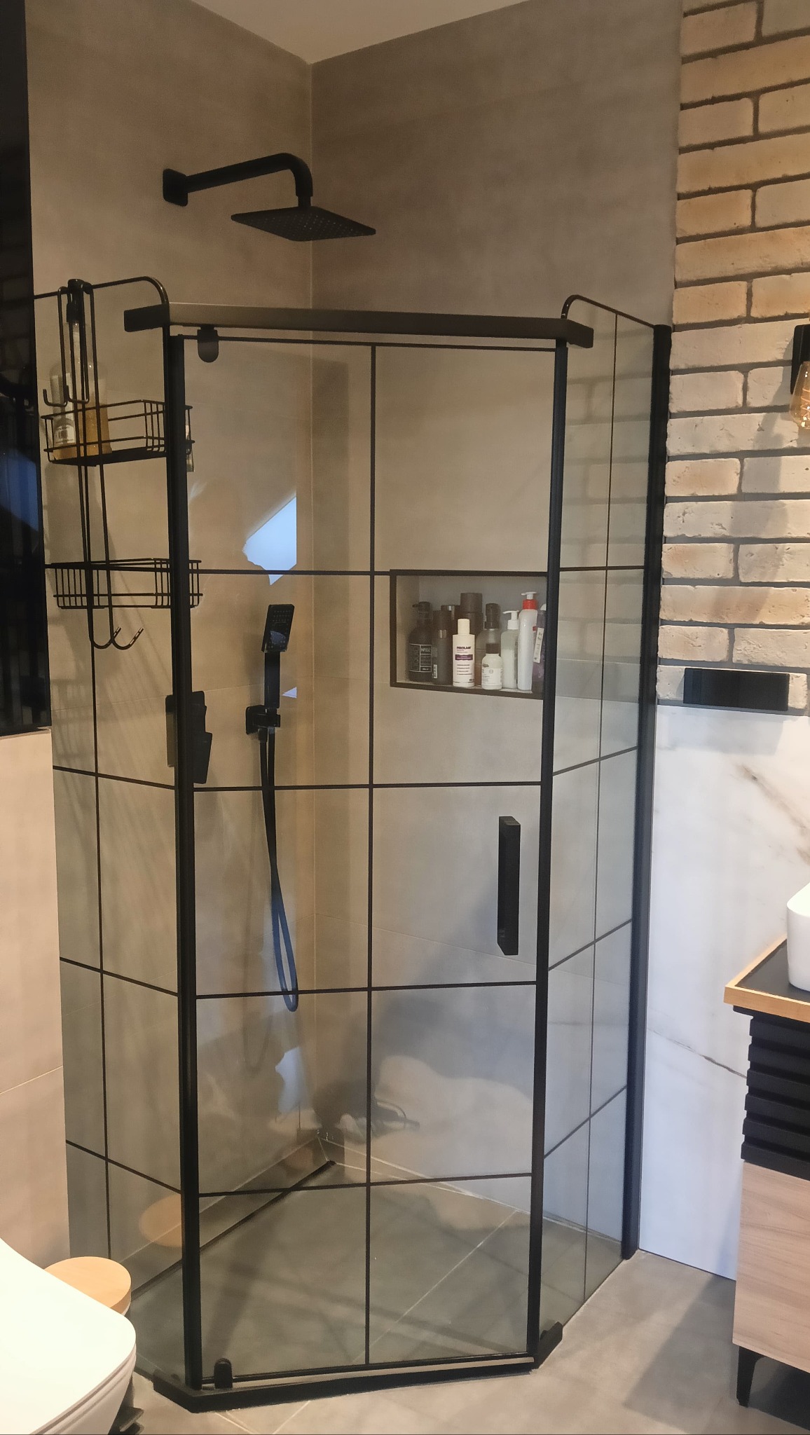 Nowoczesna kabina prysznicowa z czarnymi profilami i deszczownicą, wkomponowana w minimalistyczną łazienkę z elementami cegły i półkami na kosmetyki.