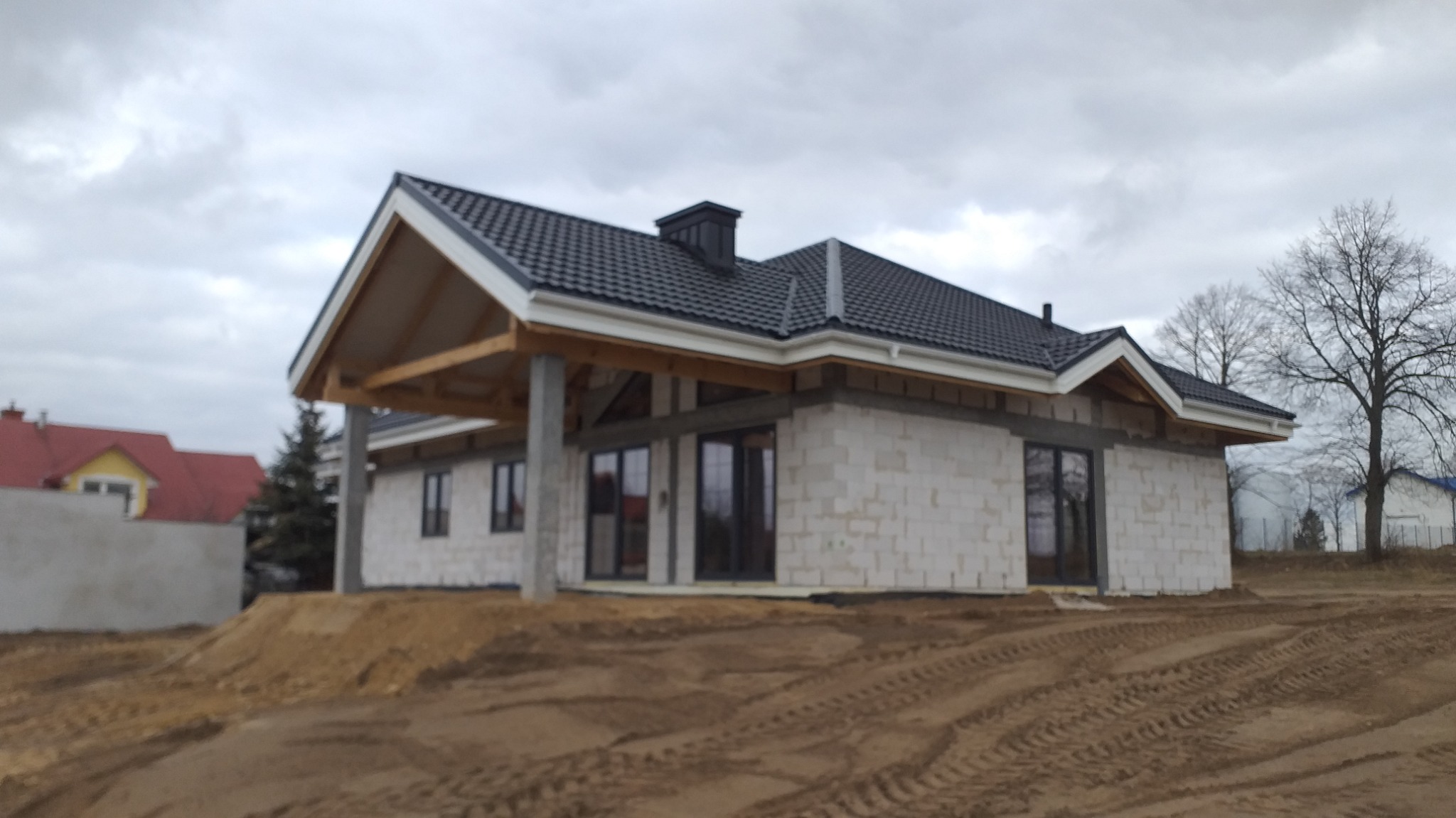 Surowy stan domu jednorodzinnego z białego bloczka, z czarną dachówką i drewnianą konstrukcją zadaszenia tarasu, na tle pochmurnego nieba i ziemi z widocznymi śladami opon.