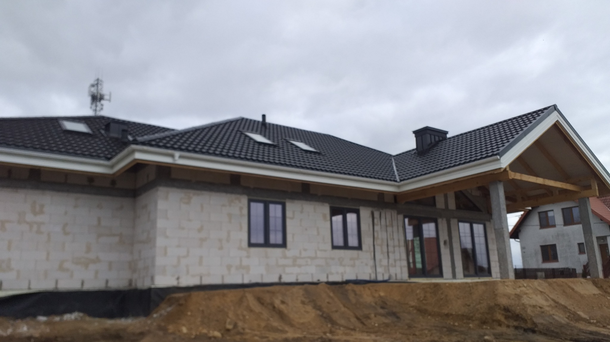 Dom jednorodzinny w trakcie budowy: ściany z betonu komórkowego, czarny dach z lukarnami, widoczne okna i drzwi. Na dachu antena. Szary dzień.
