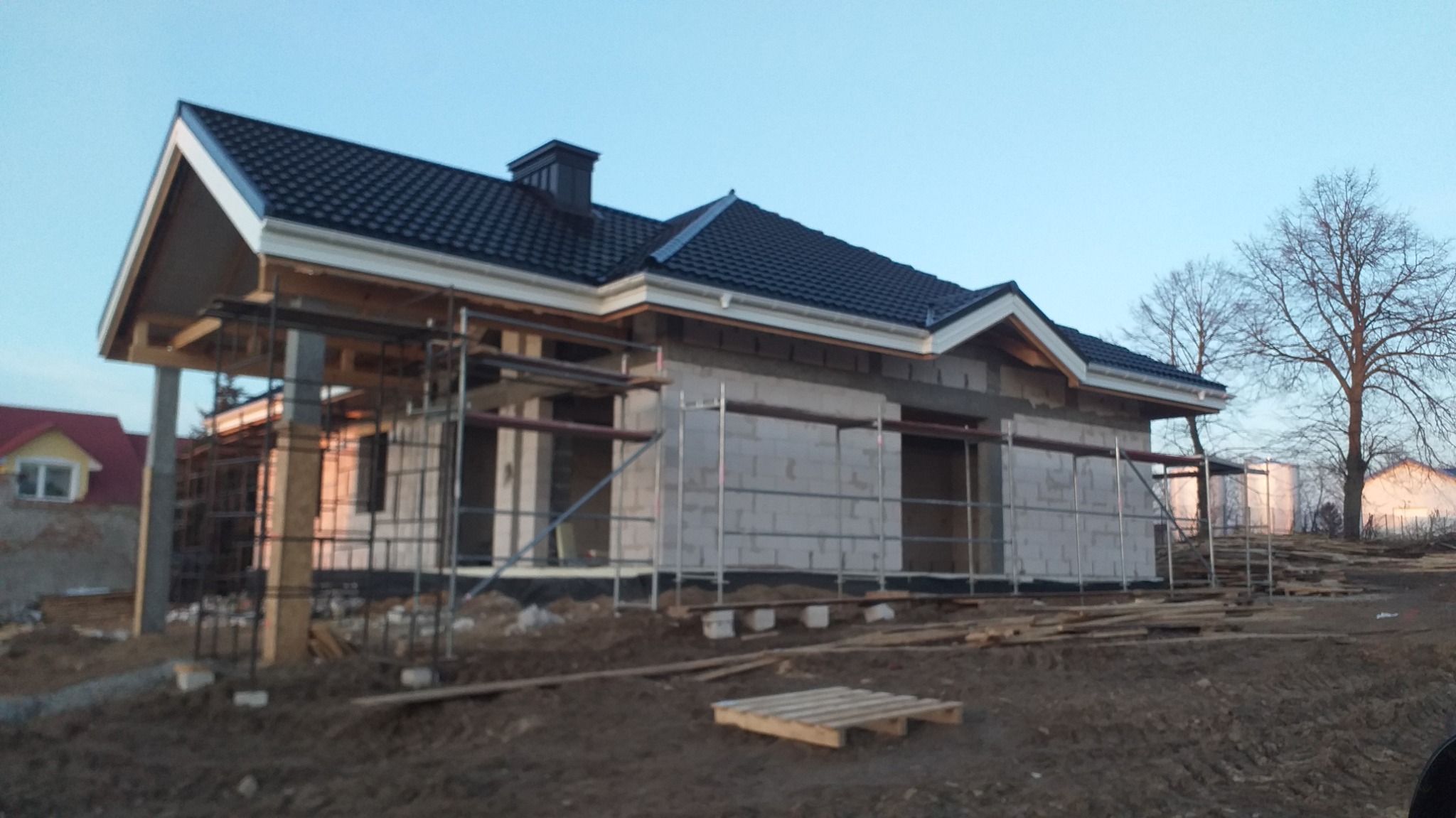 Dom w trakcie budowy z białych bloczków, z czarnym dachem i rusztowaniami. Widoczne drewniane elementy konstrukcyjne i paleta. W tle drzewo bez liści i inne budynki.