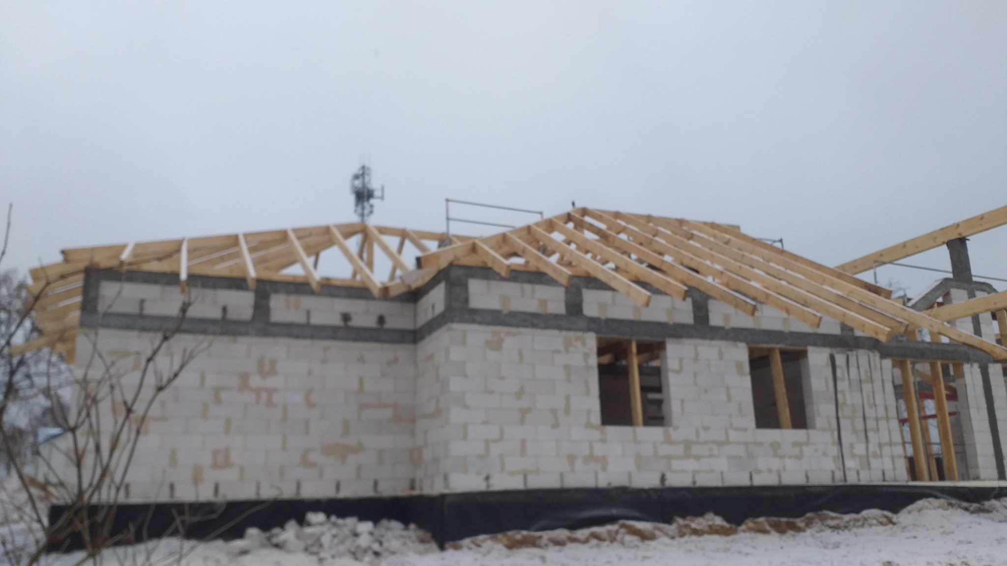 Budowa domu jednorodzinnego w stanie surowym otwartym: białe bloczki, drewniana konstrukcja dachu, okna bez szyb, zimowy krajobraz w tle.