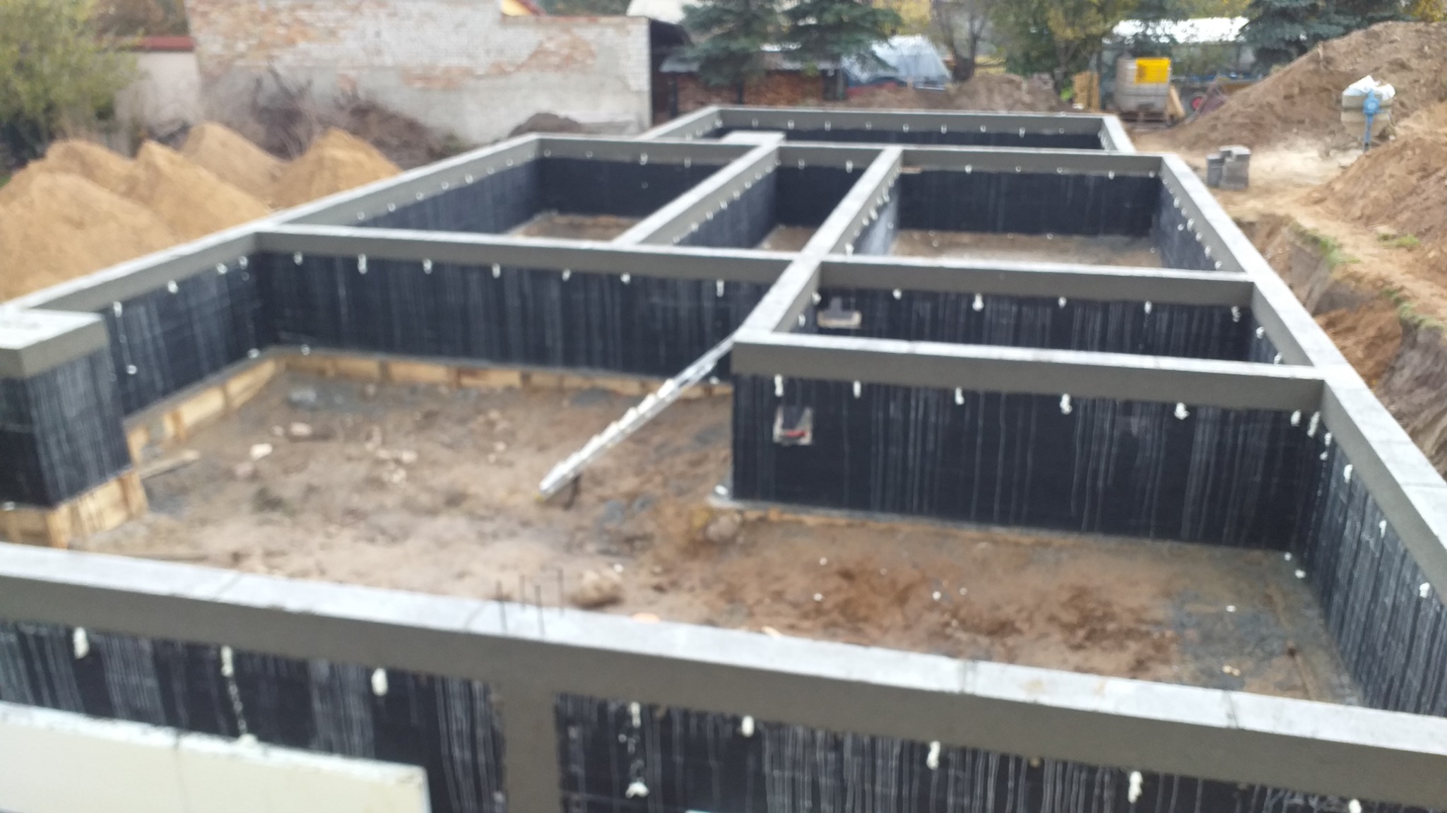 Zabezpieczone fundamenty domu jednorodzinnego z izolacją pionową i szalunkiem traconym, widok z góry na plac budowy. W tle hałdy piasku i zabudowa.