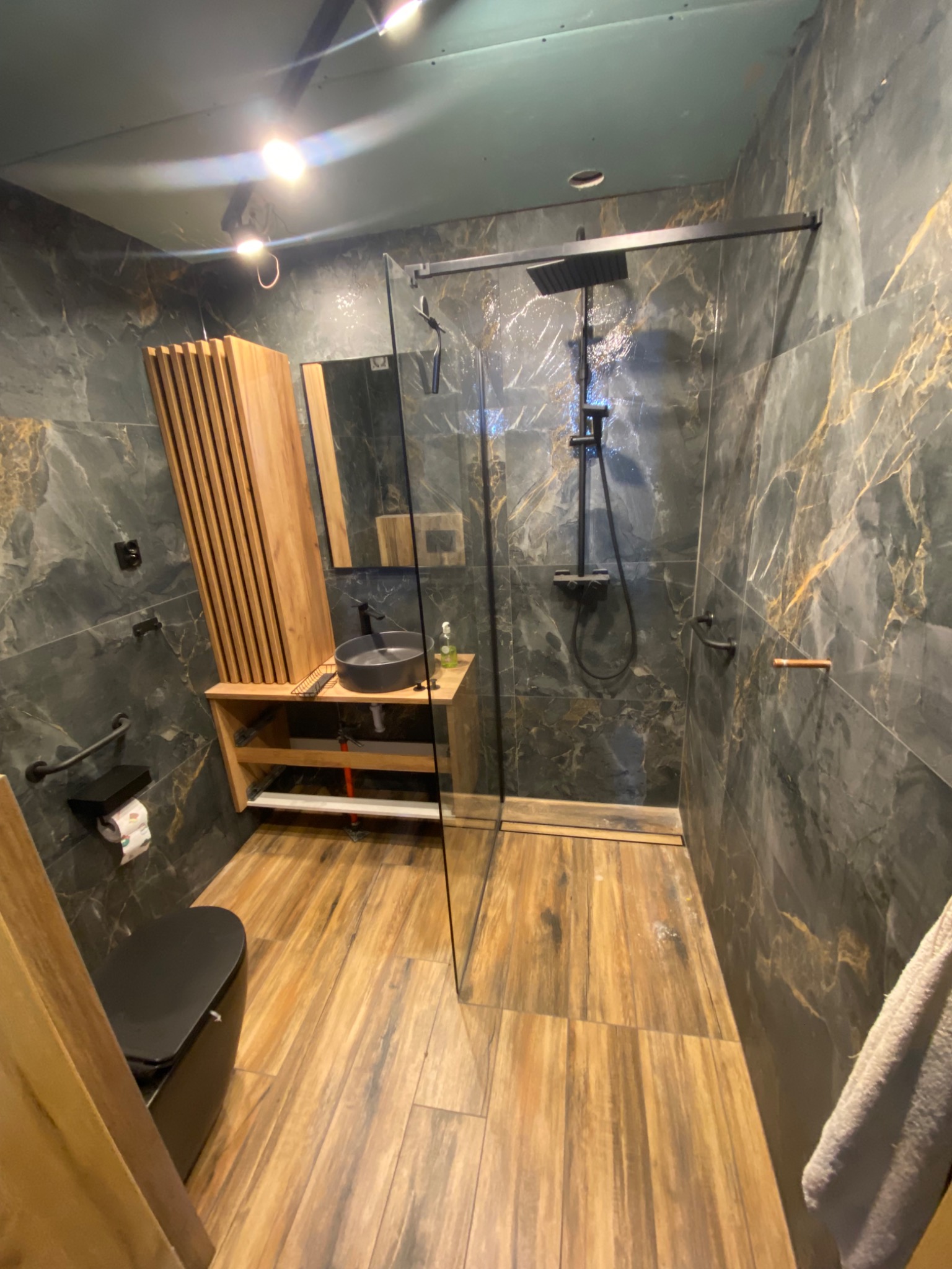 Nowoczesna łazienka z kabiną prysznicową typu walk-in, umywalką na drewnianym blacie i płytkami imitującymi marmur. Widoczne drewniane akcenty i czarna armatura.