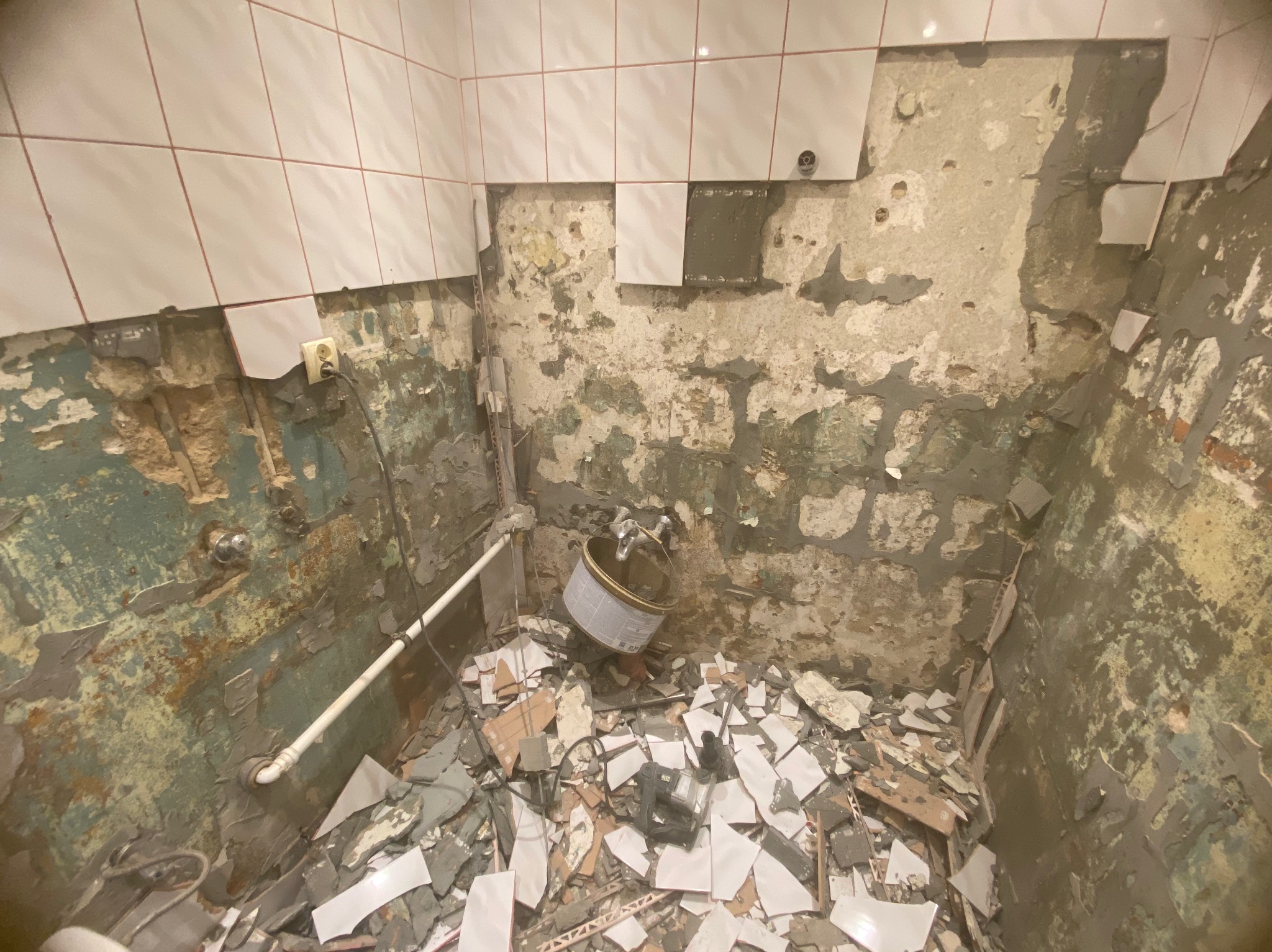 Demontaż płytek w łazience. Fragment ściany z resztkami kleju i starej farby, gruz z potłuczonych płytek na podłodze, widoczna puszka po kleju i narzędzia.