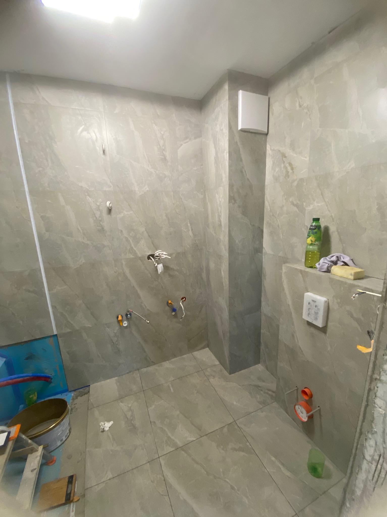Łazienka w trakcie wykańczania: szare płytki imitujące marmur, widoczne instalacje wodne, puszka z farbą i drabina. Częściowo ułożona glazura pod prysznicem.