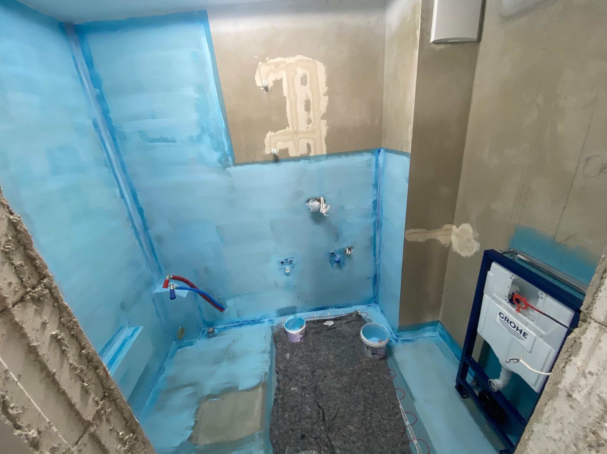 Łazienka w trakcie hydroizolacji: ściany i podłoga pokryte niebieską warstwą, widoczne instalacje, mata grzewcza i stelaż podtynkowy WC Grohe. Prace wykończeniowe.