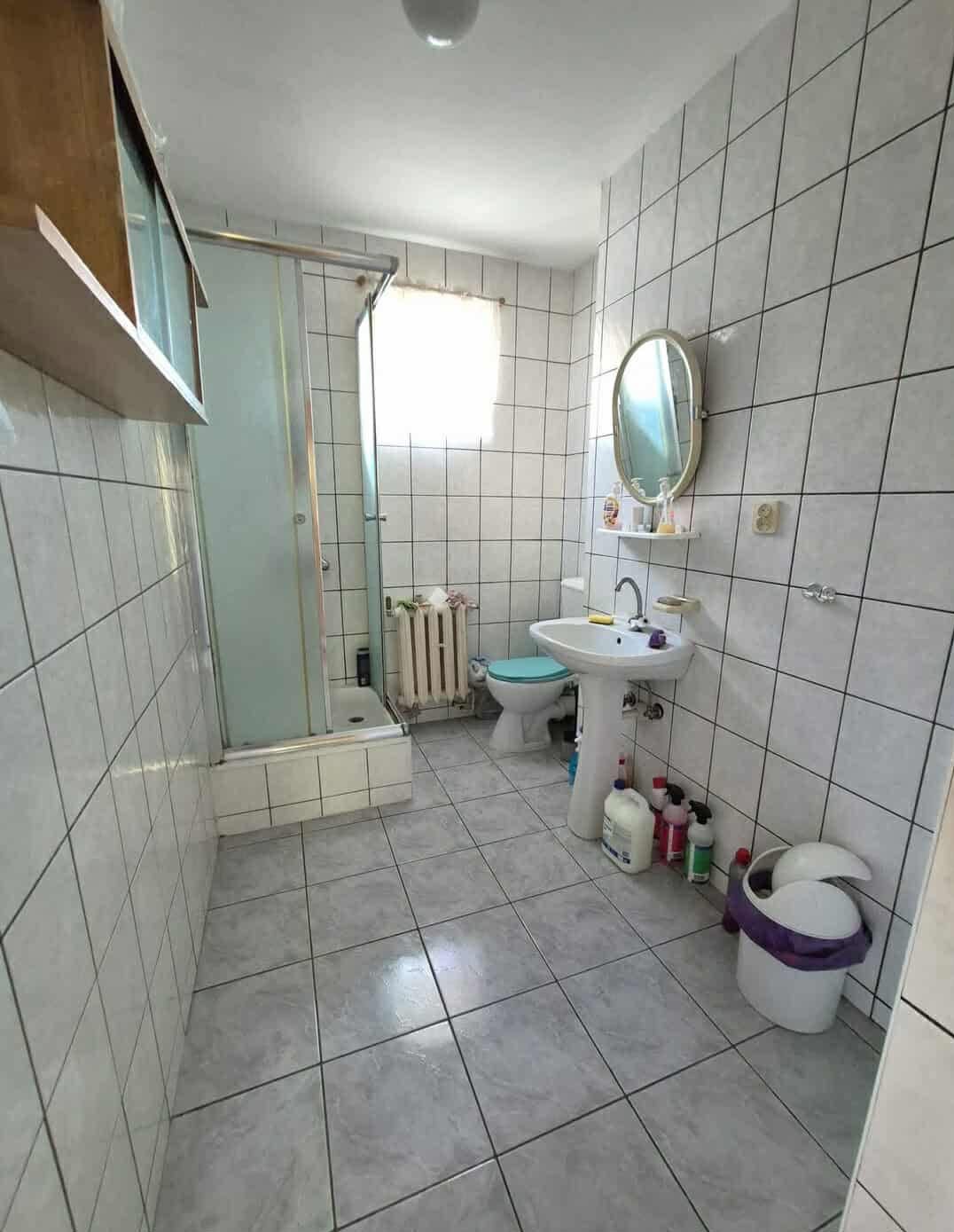 Jasna łazienka z kabiną prysznicową, umywalką na postumencie i grzejnikiem żeberkowym. Białe płytki na ścianach i podłodze. Widoczne środki czystości.