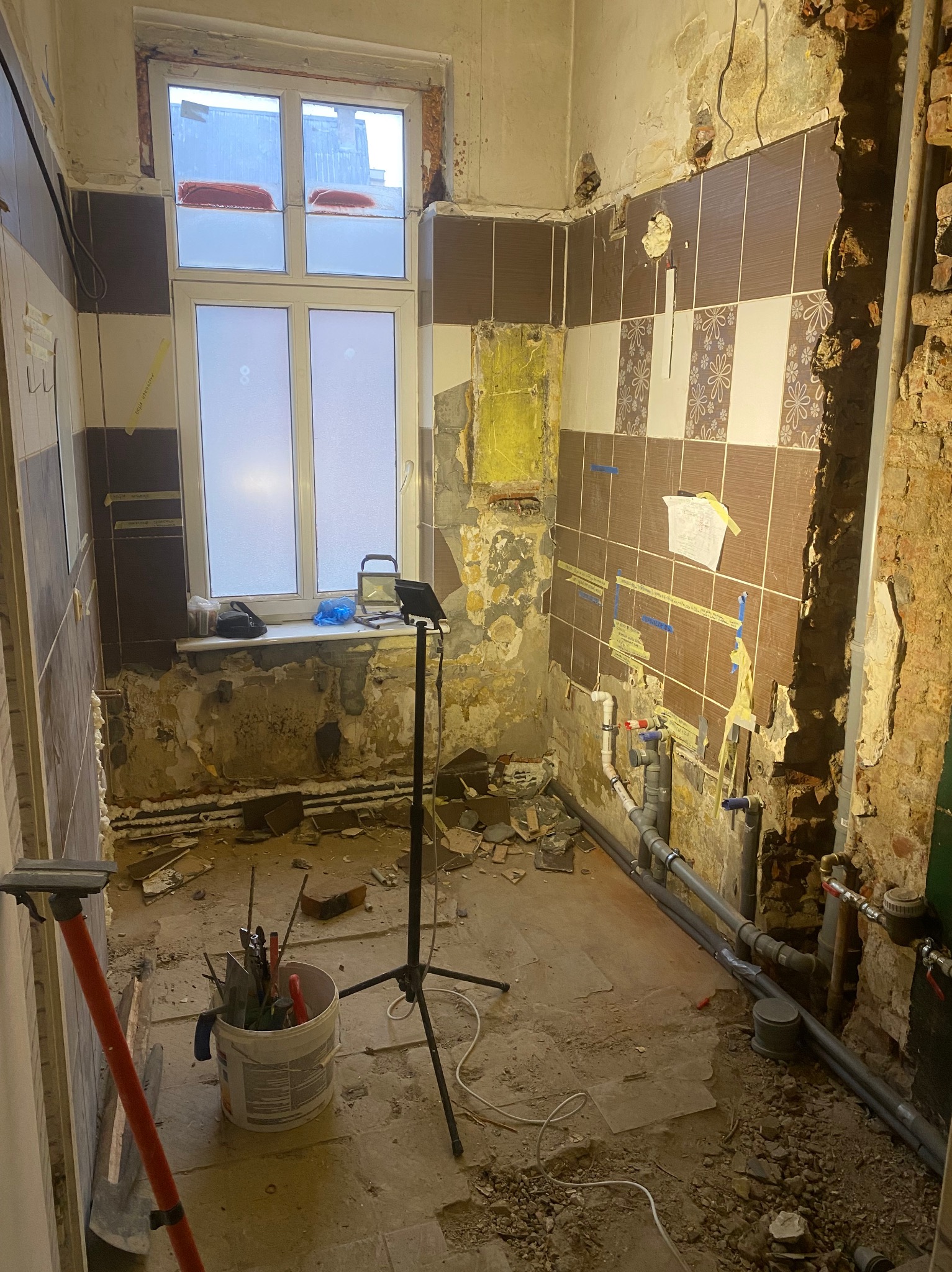 Demontaż płytek w łazience podczas remontu. Widoczne odsłonięte ściany, instalacja wodna i narzędzia w wiadrze. Okno z widokiem na zewnątrz.