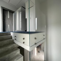 Technic Construction Dawid Gibas - Nowoczesna balustrada szklana na schodach z marmurowymi stopniami. Minimalistyczny design z metalowymi mocowaniami. W tle widoczna osoba odkurzająca podłogę.