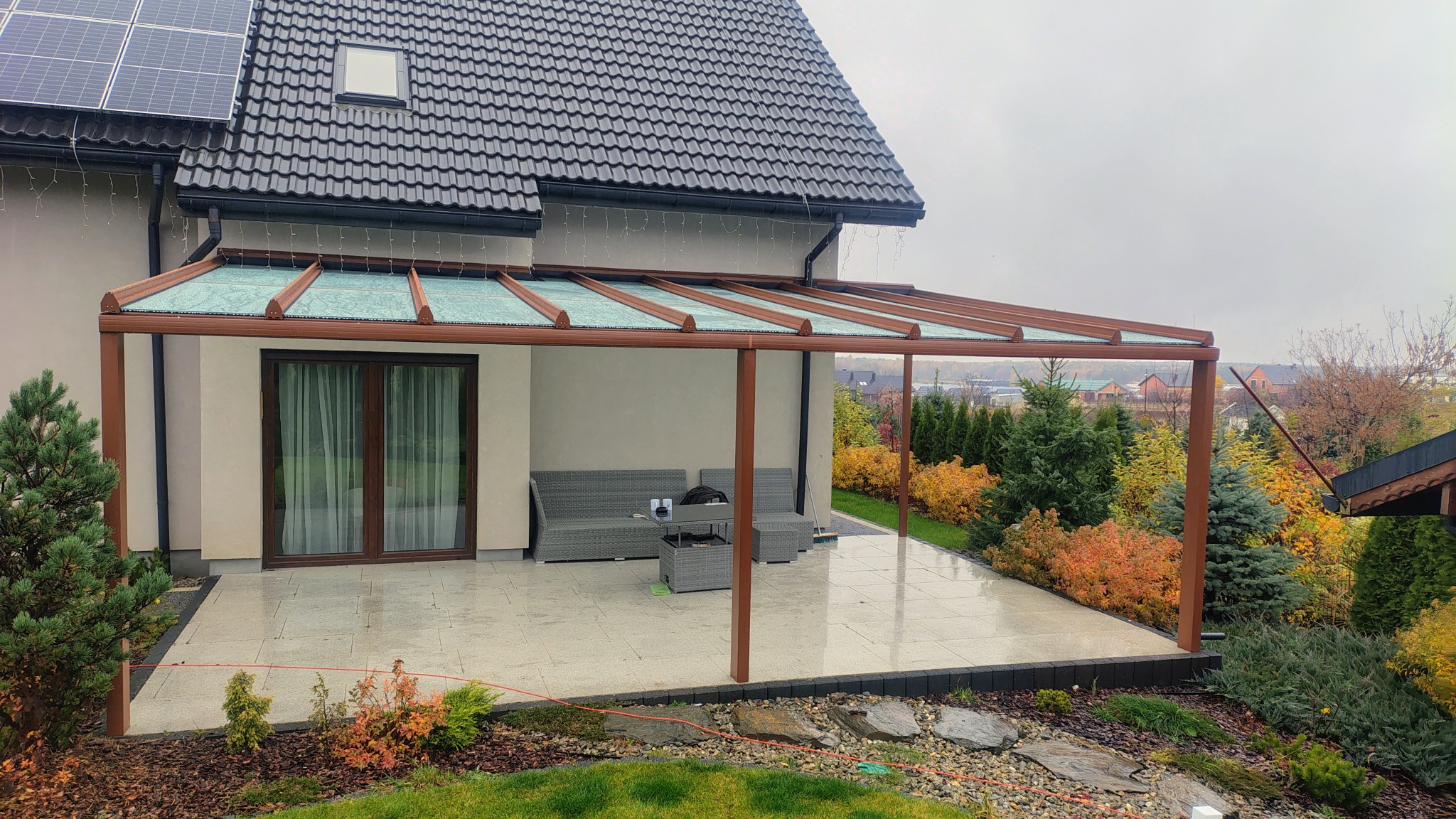 Metalowa pergola z przezroczystym zadaszeniem, brązowe słupy, szara sofa ogrodowa i stolik na jasnym tarasie z kamiennych płyt przy domu z panelami słonecznymi.