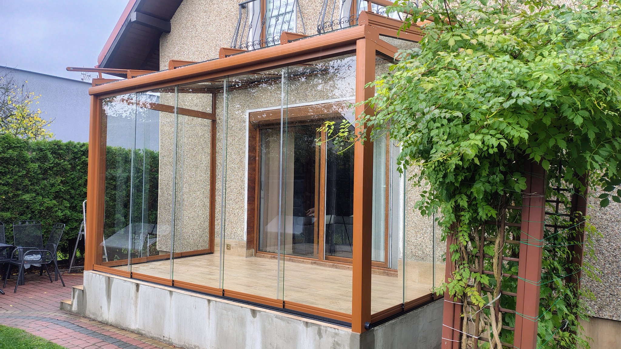Zadaszony taras z przesuwnymi panelami szklanymi i drewnianą konstrukcją, widok z zewnątrz, zieleń w tle, jasne płytki na podłodze.