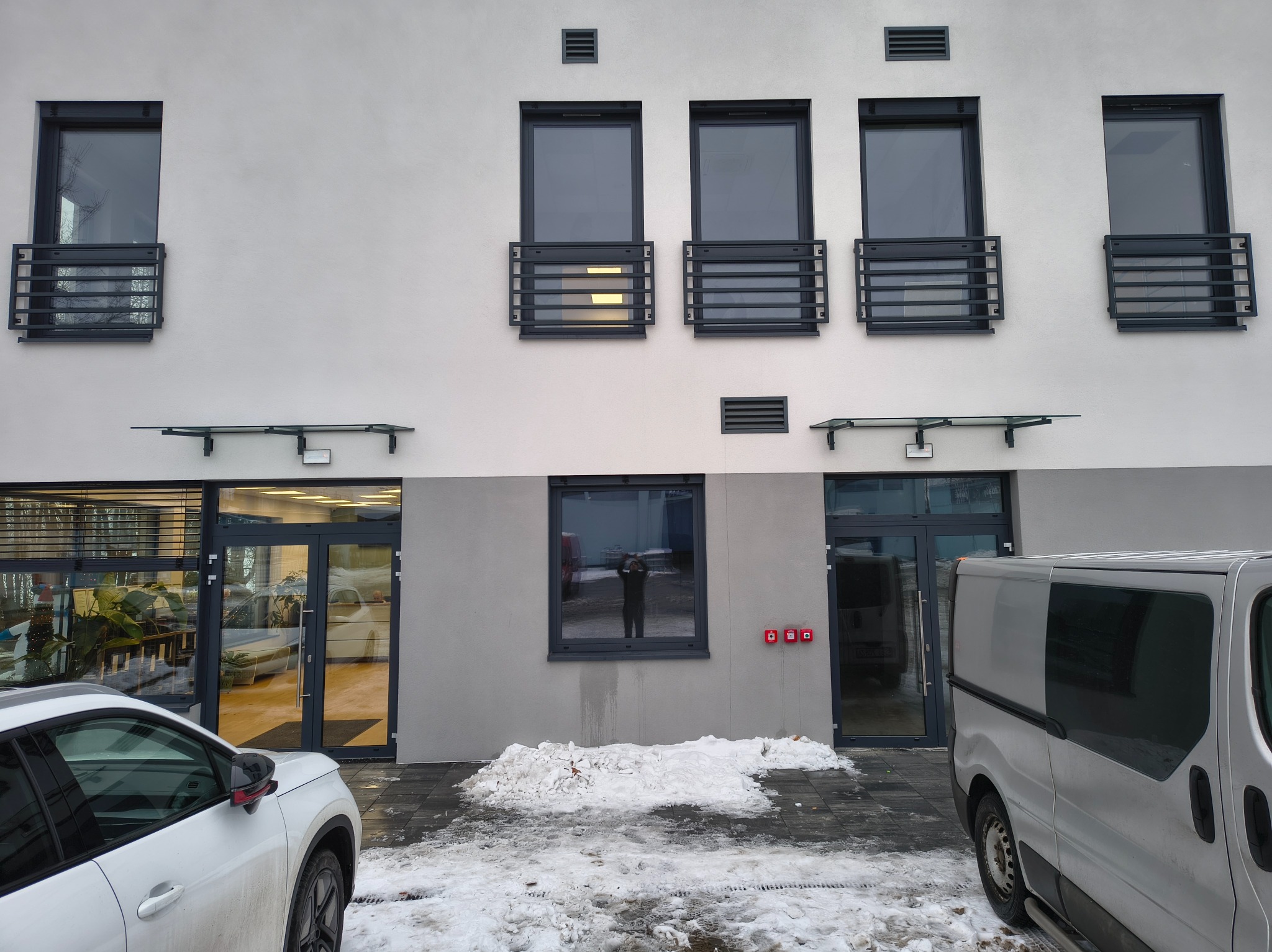 Elewacja budynku z oknami zabezpieczonymi metalowymi balustradami. Widoczne wejście z przeszklonymi drzwiami i zaparkowane samochody. Zima, śnieg.