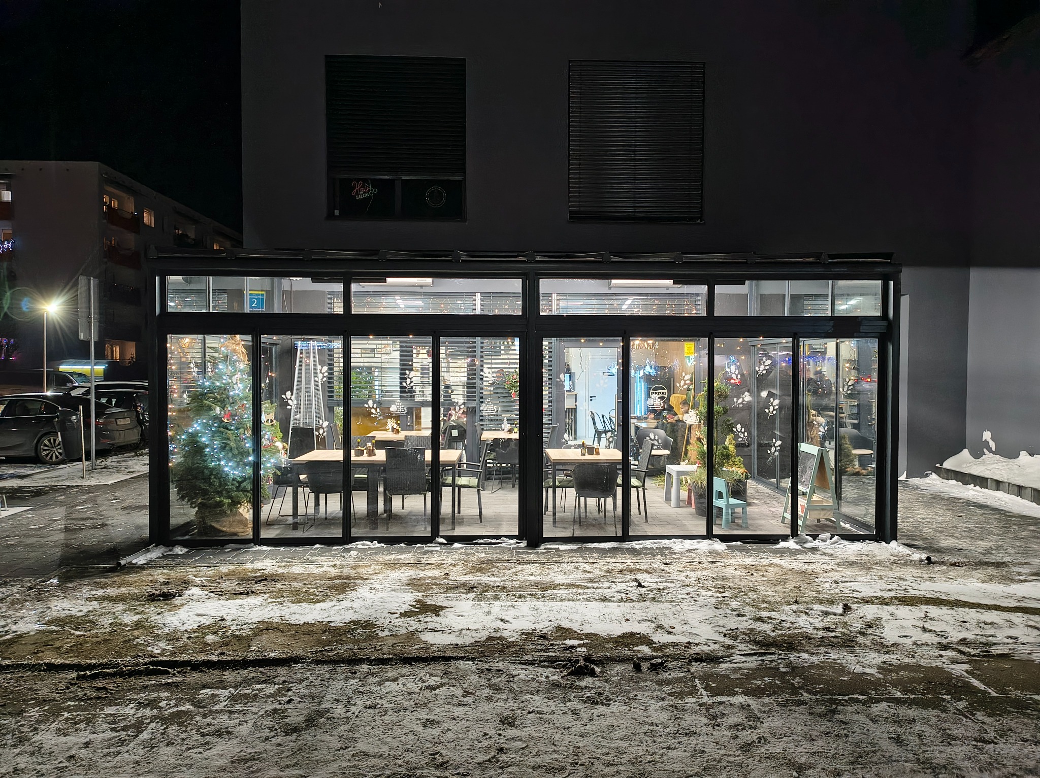 Zimowa zabudowa szklana lokalu gastronomicznego z widocznym wnętrzem: stoliki, krzesła, dekoracje świąteczne. Na zewnątrz śnieg i lód.