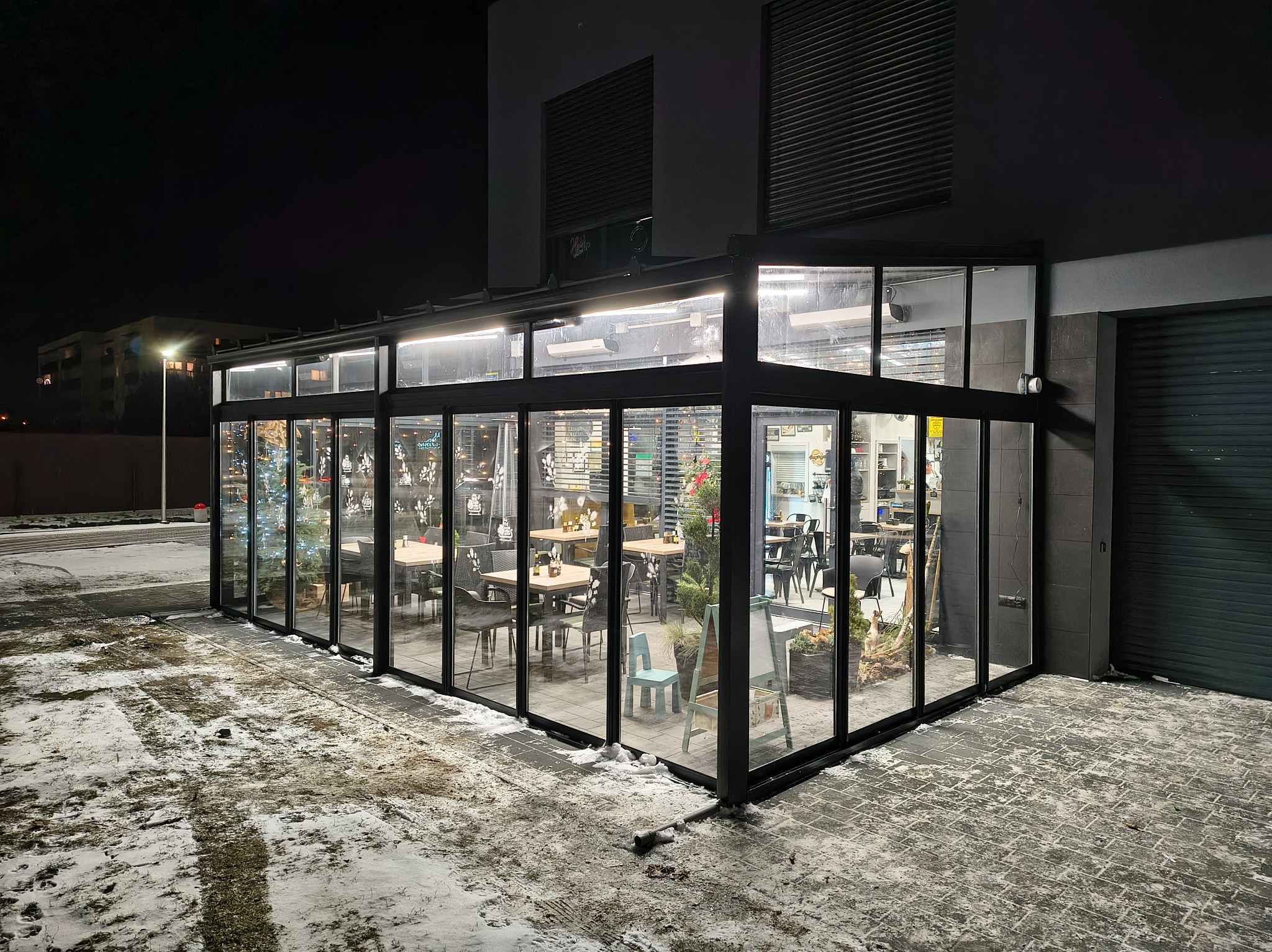 Szklana zabudowa tarasu restauracji z czarnymi ramami, widoczna z zewnątrz w zimowej scenerii. Wnętrze oświetlone, widać stoliki i krzesła.