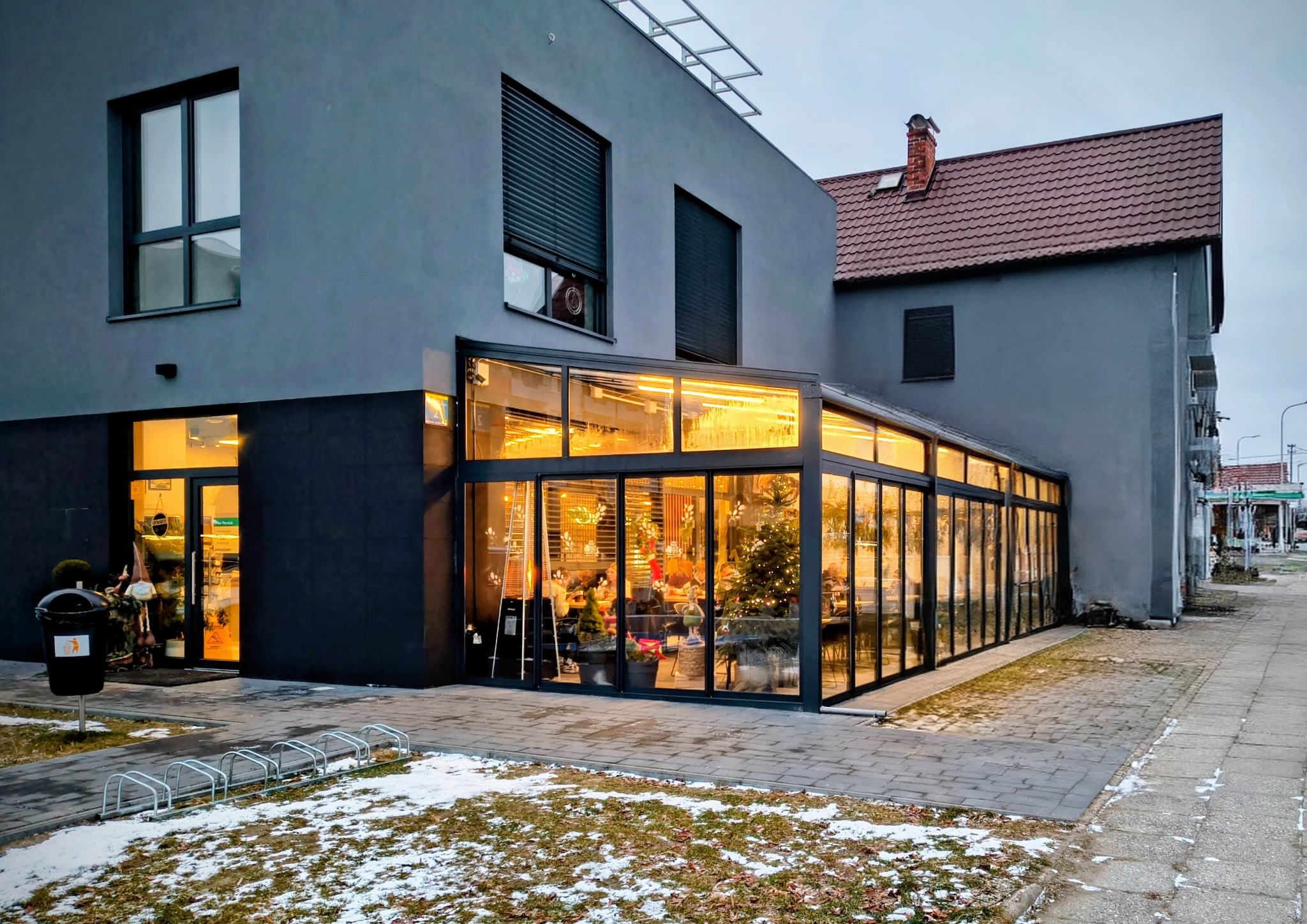 Szklana zabudowa tarasu przy budynku z szarą elewacją, widoczna z zewnątrz. Wewnątrz oświetlenie i dekoracje świąteczne. Na zewnątrz chodnik i resztki śniegu.