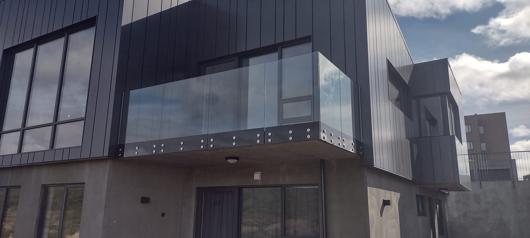 Nowoczesny budynek z balkonem zabezpieczonym szklaną balustradą mocowaną punktowo. Elewacja w odcieniach szarości, minimalistyczny design.