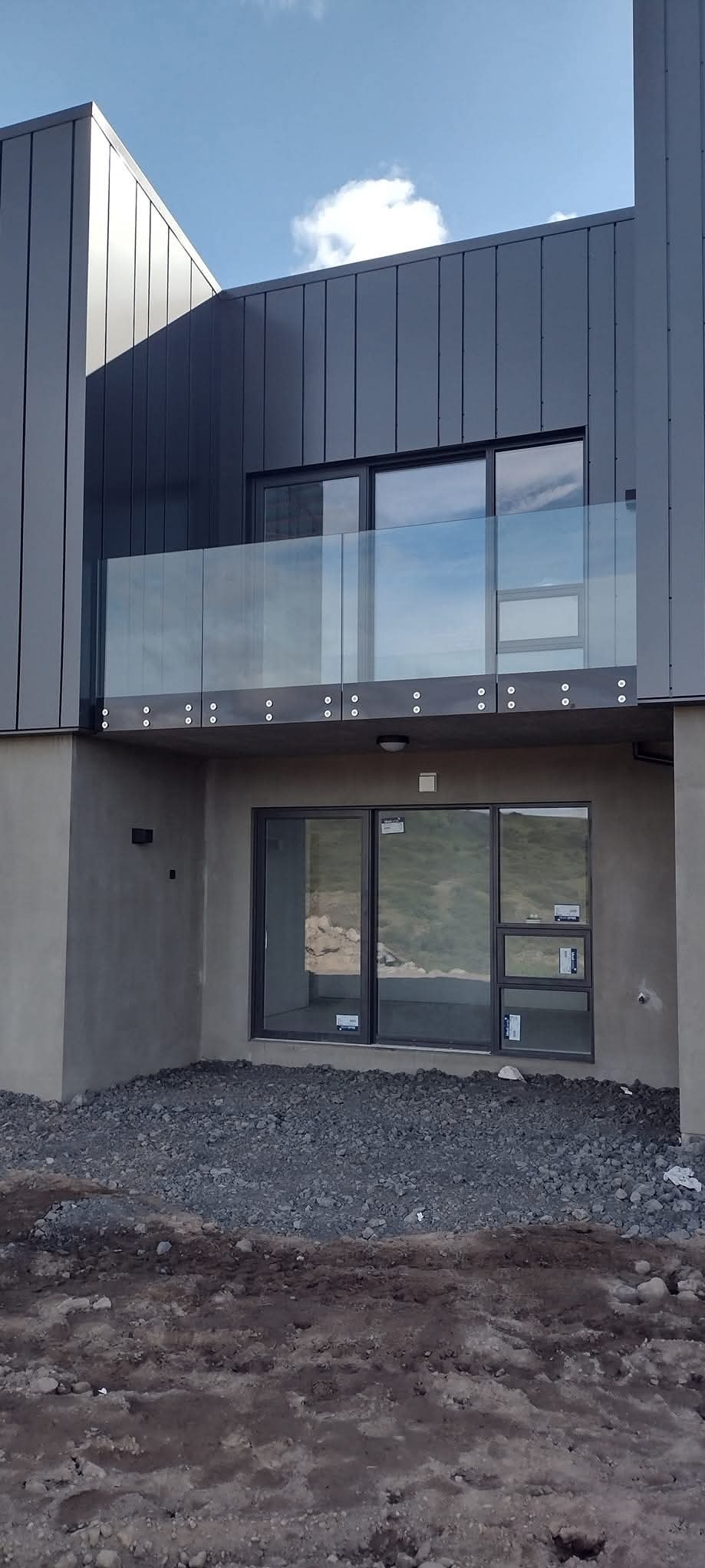 Nowoczesny budynek z balkonem zabezpieczonym szklaną balustradą mocowaną punktowo. Widoczne szare elewacje i okna. Teren wokół budynku w trakcie prac wykończeniowych.