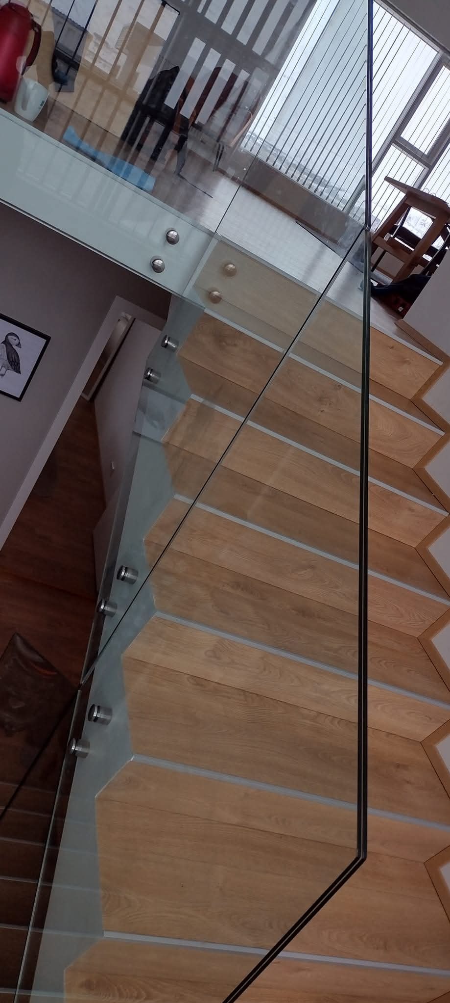 Szklana balustrada z drewnianymi schodami, widok z góry. Elementy mocujące ze stali nierdzewnej. W tle widoczne okno z żaluzjami i fragment pomieszczenia.