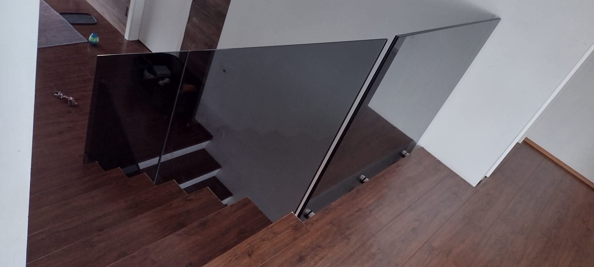 Szklana balustrada schodowa z ciemnego szkła, mocowana punktowo do drewnianych schodów. Widok z góry, ukazujący detale mocowania i przejrzystość szkła.