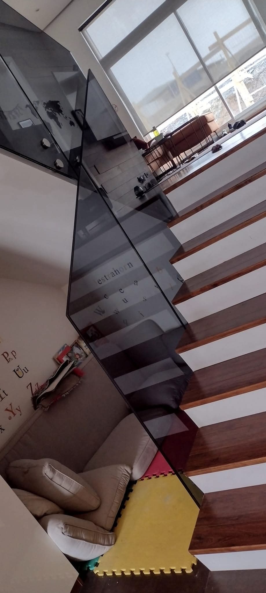 Szklana balustrada schodowa z mocowaniami punktowymi, widok z dołu na drewniane stopnie i fragment pokoju dziecięcego z kanapą i puzzlami piankowymi.