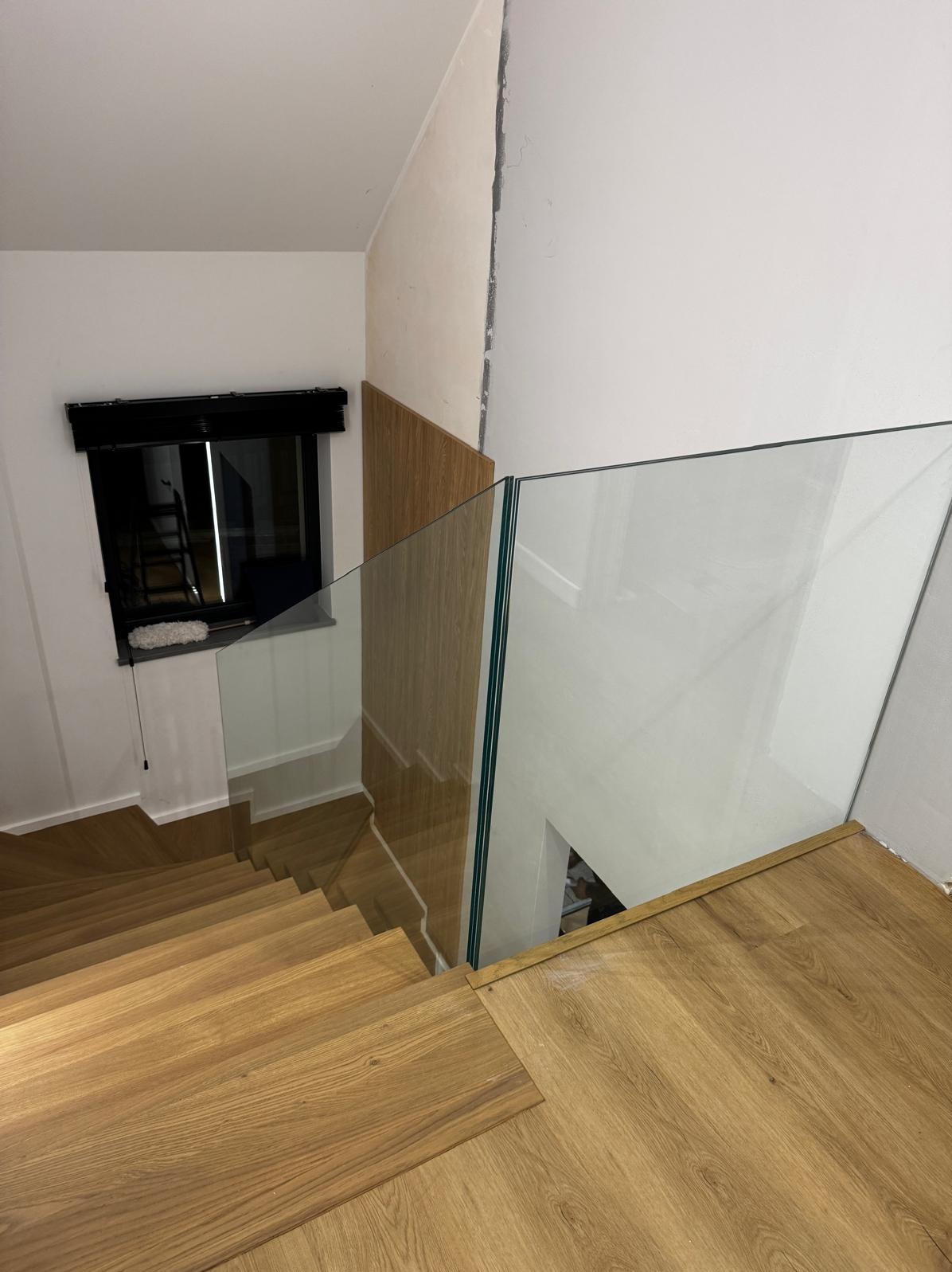 Szklana balustrada zabezpiecza drewniane schody w jasnym wnętrzu. Widok z góry ukazuje detale montażu i minimalistyczny design.