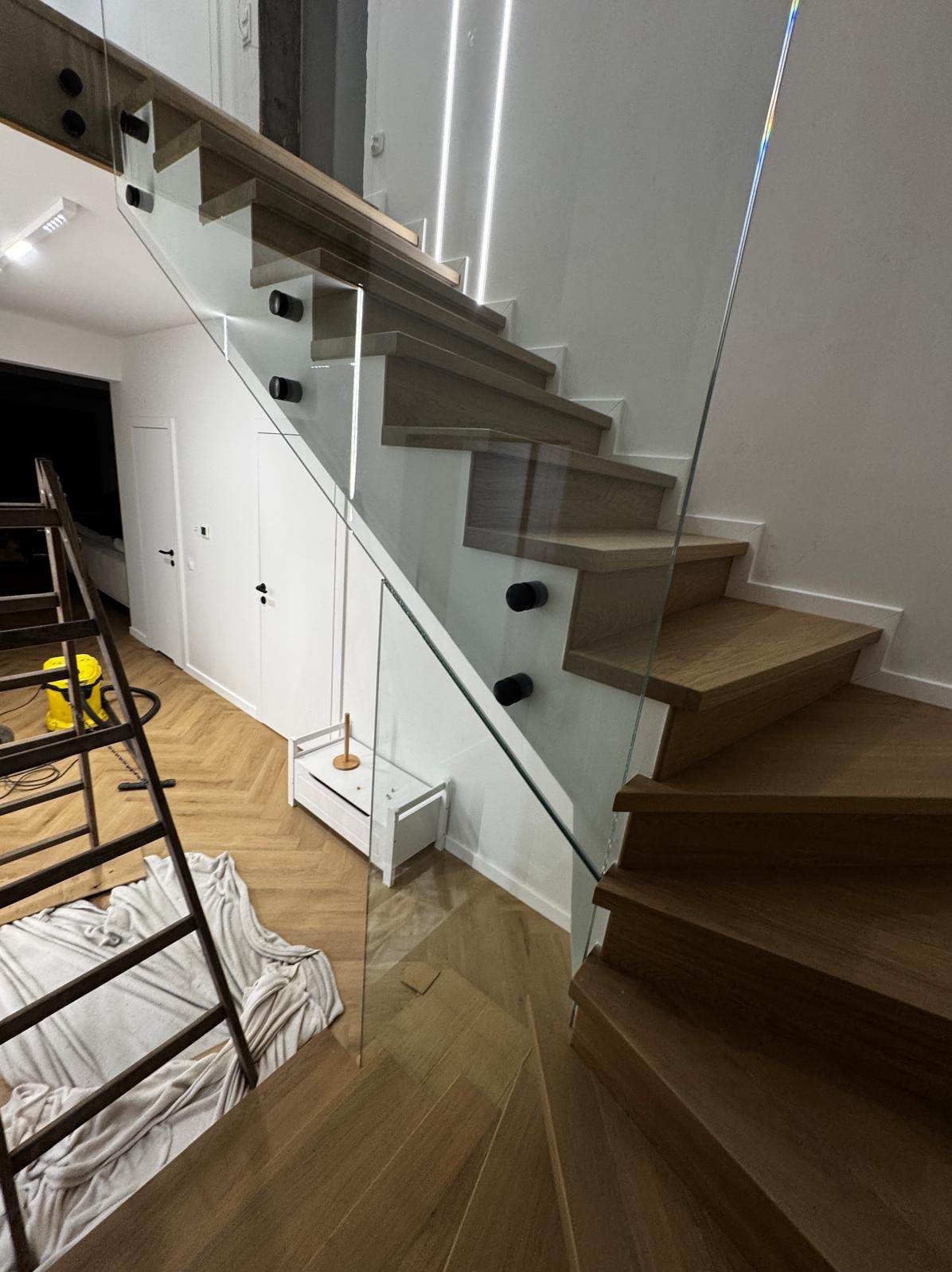 Drewniane schody z balustradą ze szkła mocowaną na czarnych uchwytach. Widoczna drabina i akcesoria montażowe na parkiecie w jodełkę.