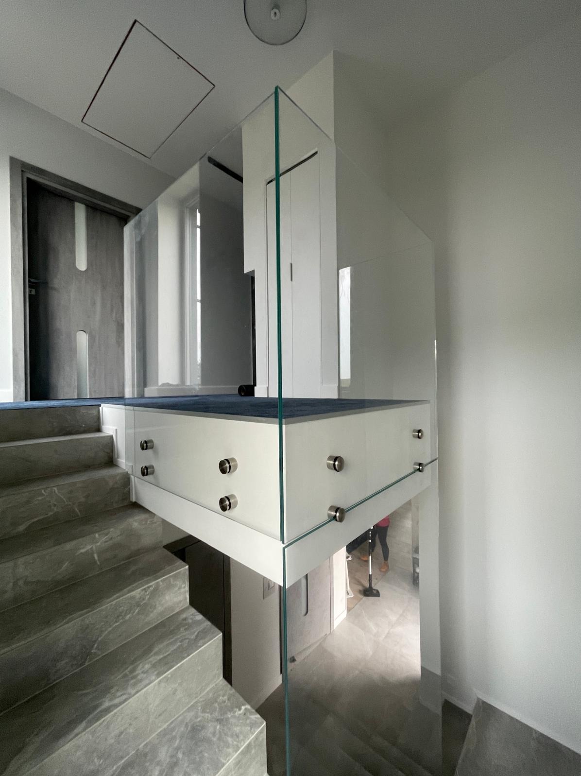 Nowoczesna balustrada szklana na schodach z marmurowymi stopniami. Minimalistyczny design z metalowymi mocowaniami. W tle widoczna osoba odkurzająca podłogę.