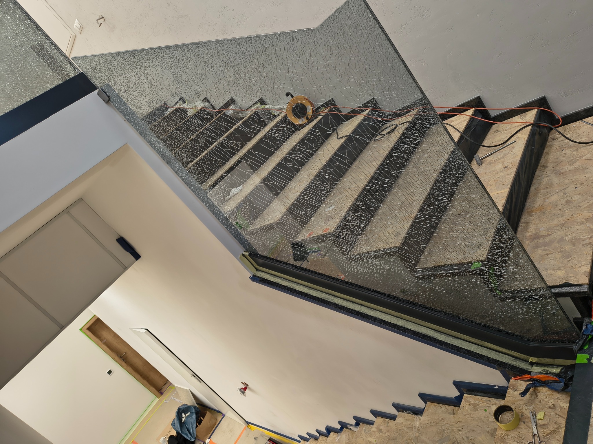 Szklana balustrada schodowa z widoczną strukturą szkła, zamontowana na drewnianych stopniach w trakcie prac wykończeniowych. Widoczne narzędzia i przewody.