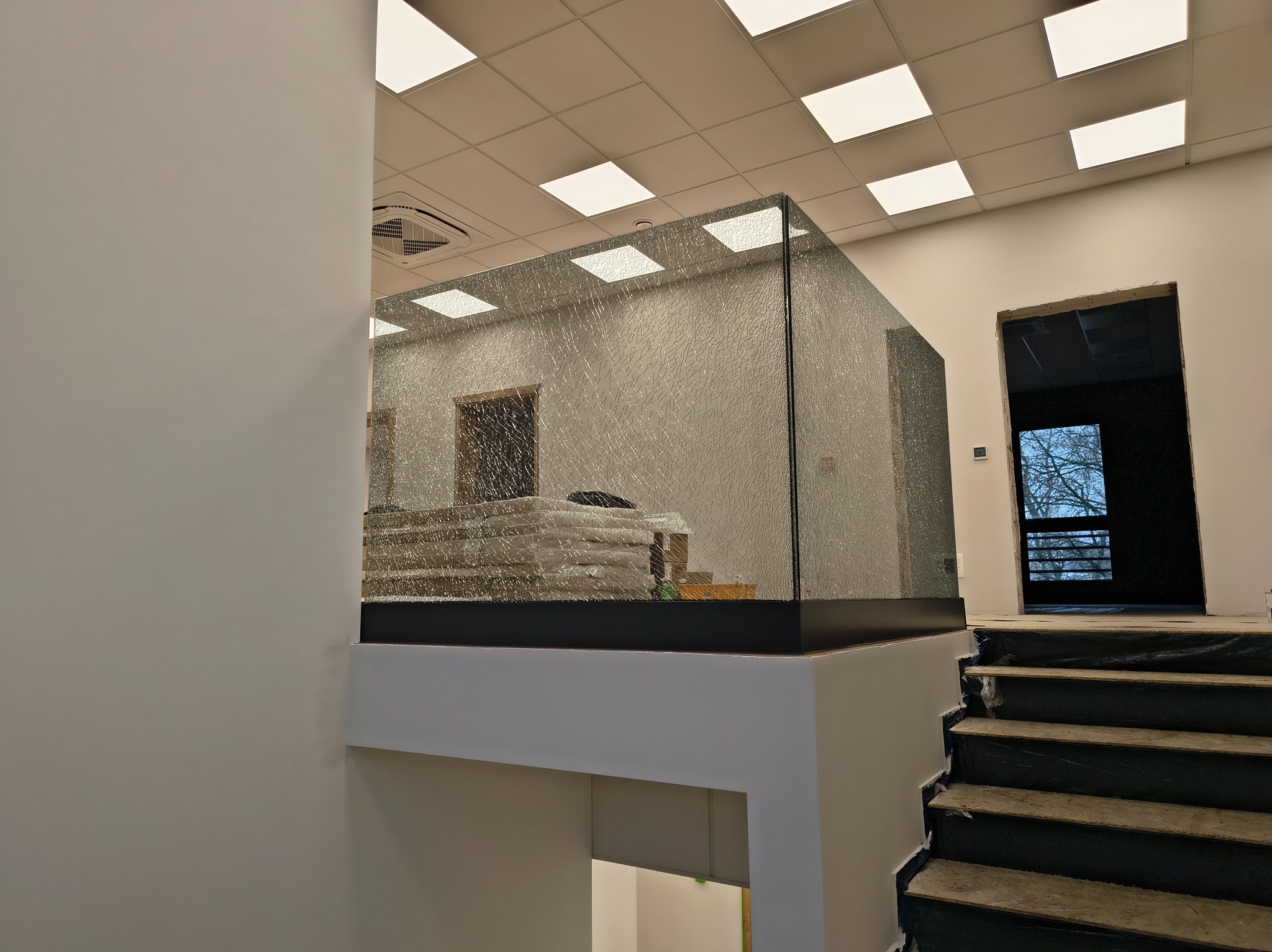 Szklana balustrada z efektem pęknięć na antresoli, widok z dołu schodów. W tle widoczne materiały budowlane i otwór drzwiowy.