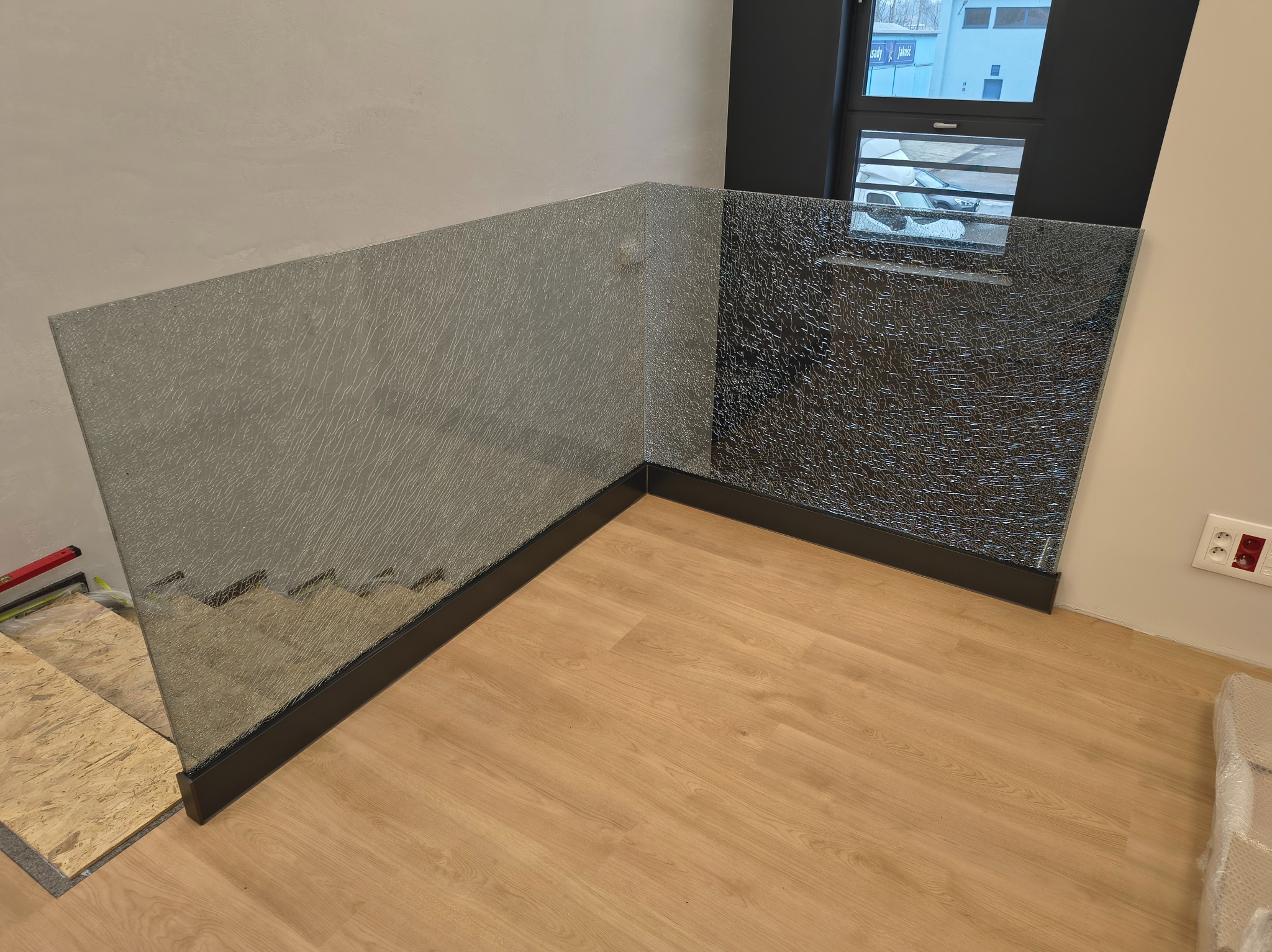 Szklana balustrada z czarną ramą, zamontowana w rogu pomieszczenia. Dwie tafle szkła o różnej fakturze, jedna gładka, druga z efektem spękania. Widoczna podłoga z paneli.