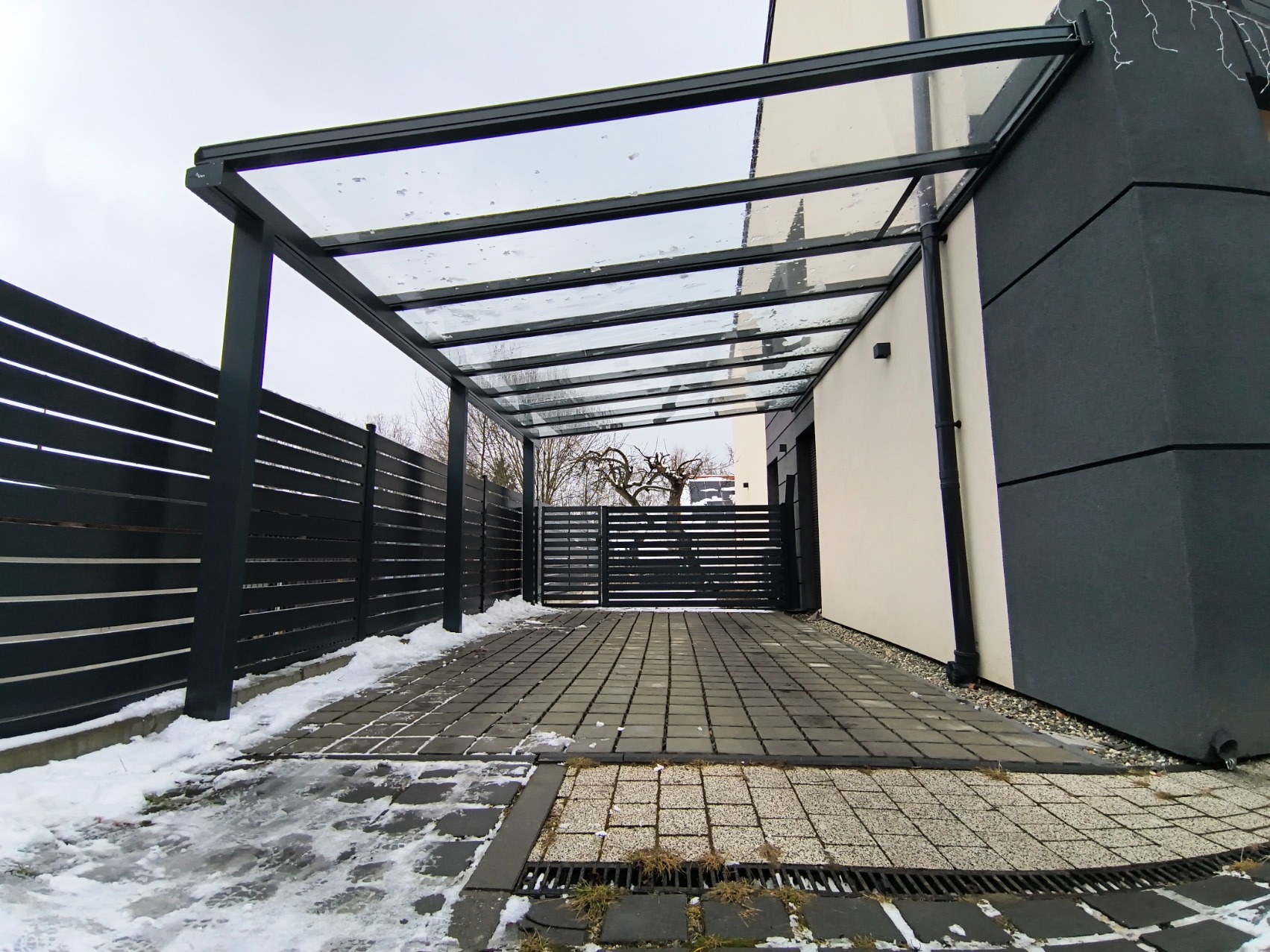 Nowoczesna, przeszklona pergola z czarną konstrukcją, przylegająca do budynku. Widoczny fragment posesji z kostką brukową i ogrodzeniem w zimowej scenerii.