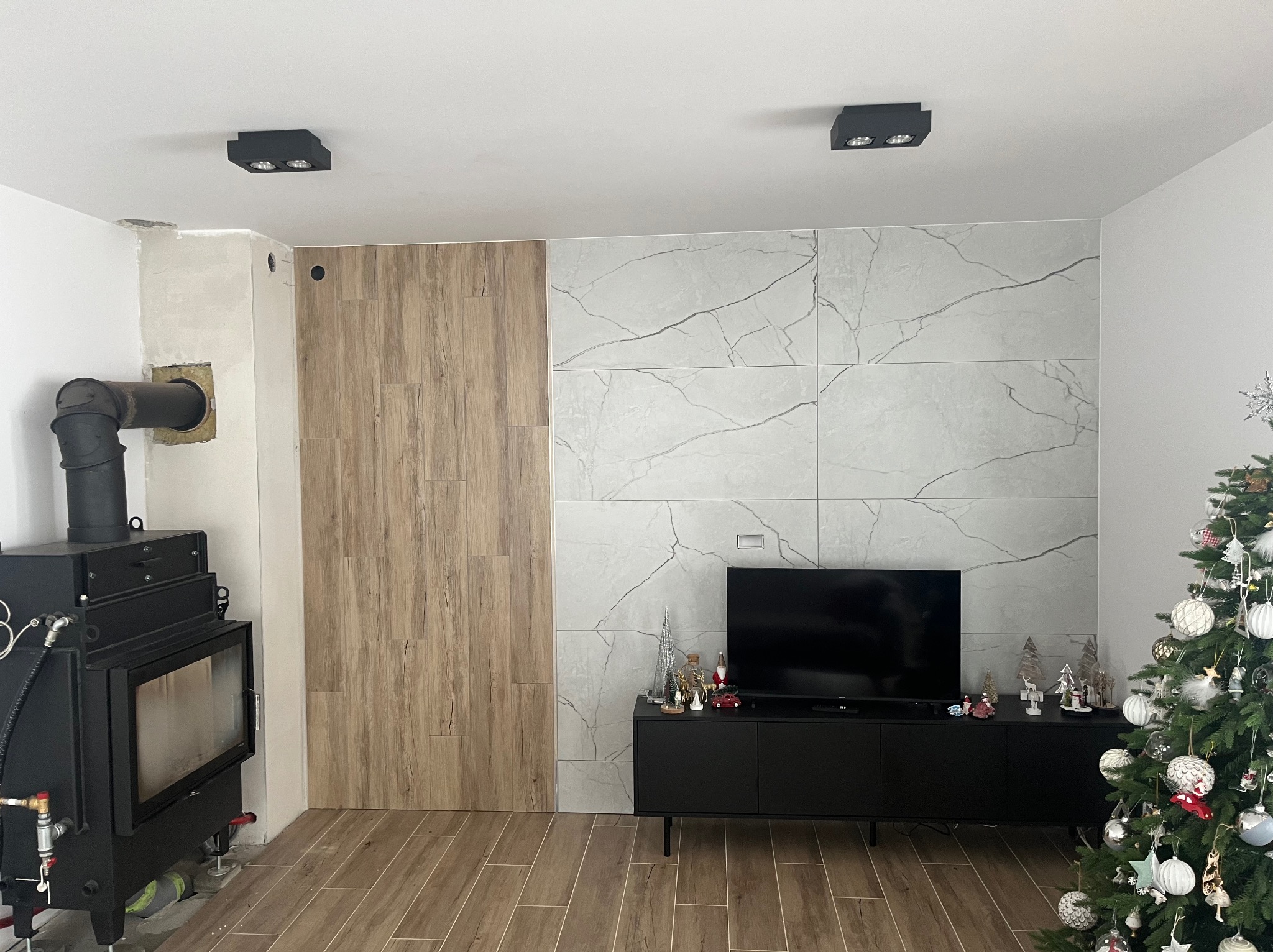 Salon po remoncie: ściana z płytek imitujących drewno i marmur, czarny kominek i szafka RTV, podłoga z paneli. Świąteczny wystrój z choinką.