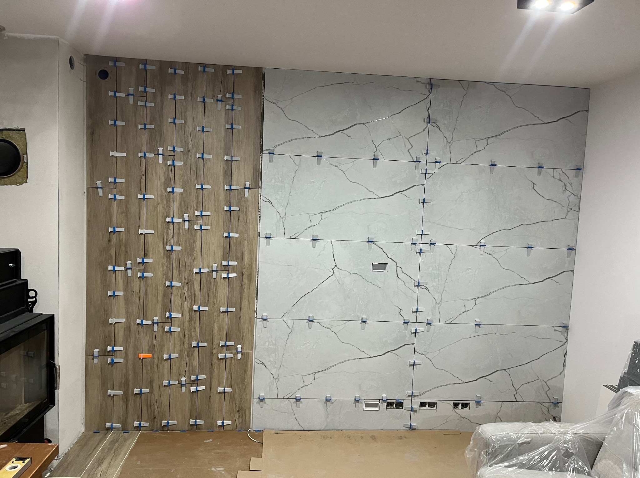 Fragment ściany z częściowo ułożonymi płytkami imitującymi drewno i marmur, z użyciem klipsów poziomujących. Widoczne otwory na gniazdka elektryczne.