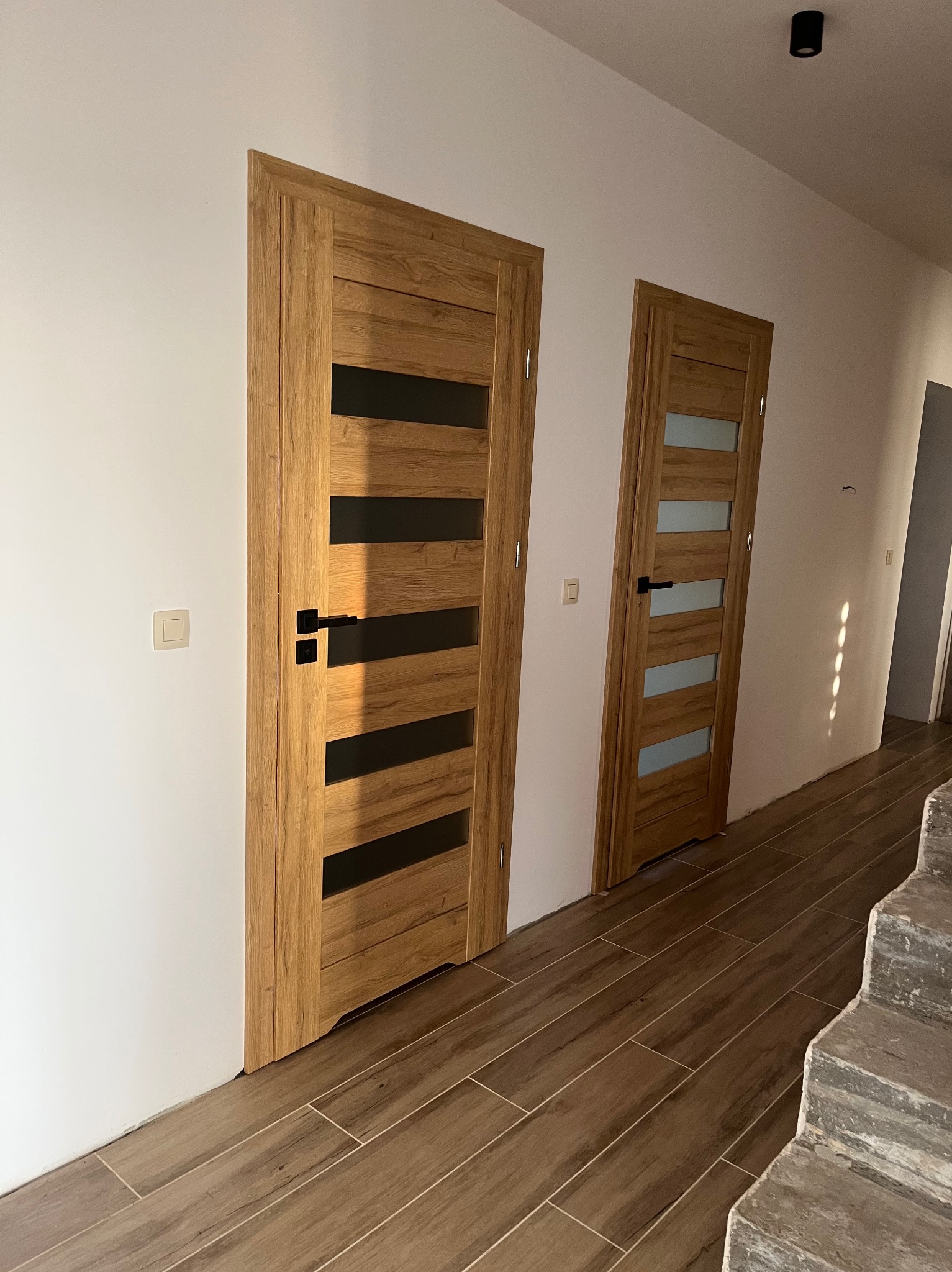 Dwa drewniane drzwi wewnętrzne z czarnymi i przezroczystymi wstawkami, zamontowane w jasnym korytarzu z podłogą imitującą drewno. Widoczne schody w stanie surowym.
