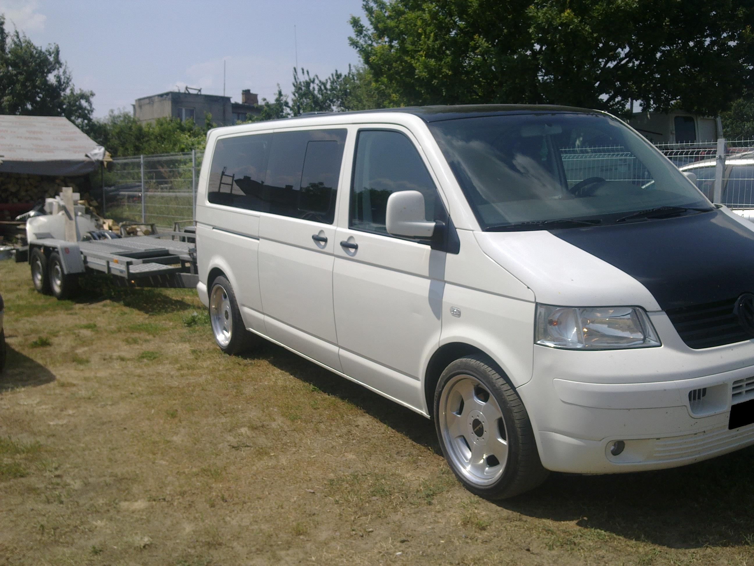 Biały bus z czarną maską i dachem, ciągnący dwuosiową przyczepę lawetę na trawiastym terenie.