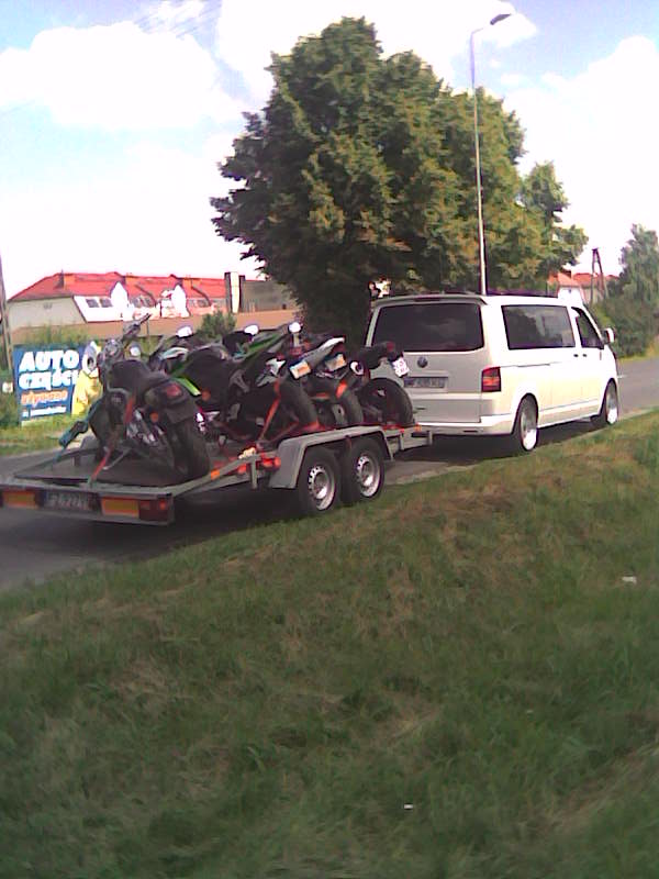 Biały bus z przyczepą przewożącą kilka motocykli, zaparkowany na poboczu, w tle budynki i drzewo.