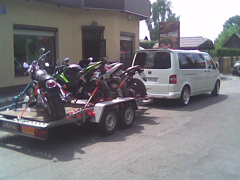 Biały van Volkswagen ciągnie przyczepę z kilkoma motocyklami na tle budynku z szyldem piekarni.