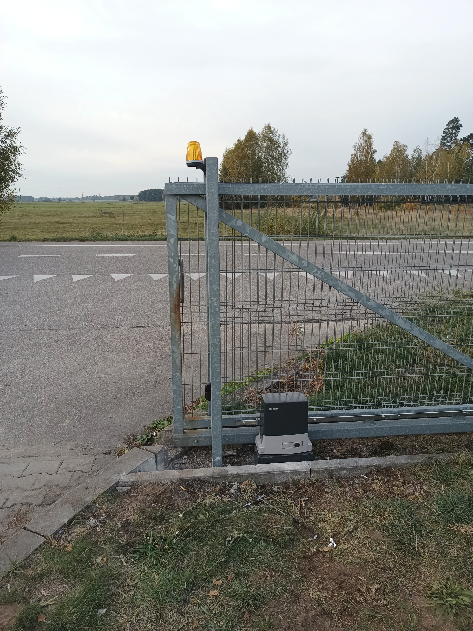 Otwarta brama panelowa z automatyką Beninca i lampą ostrzegawczą, widok z boku na tle drogi i łąki. Szara kolorystyka bramy i otoczenia, pochmurne niebo.